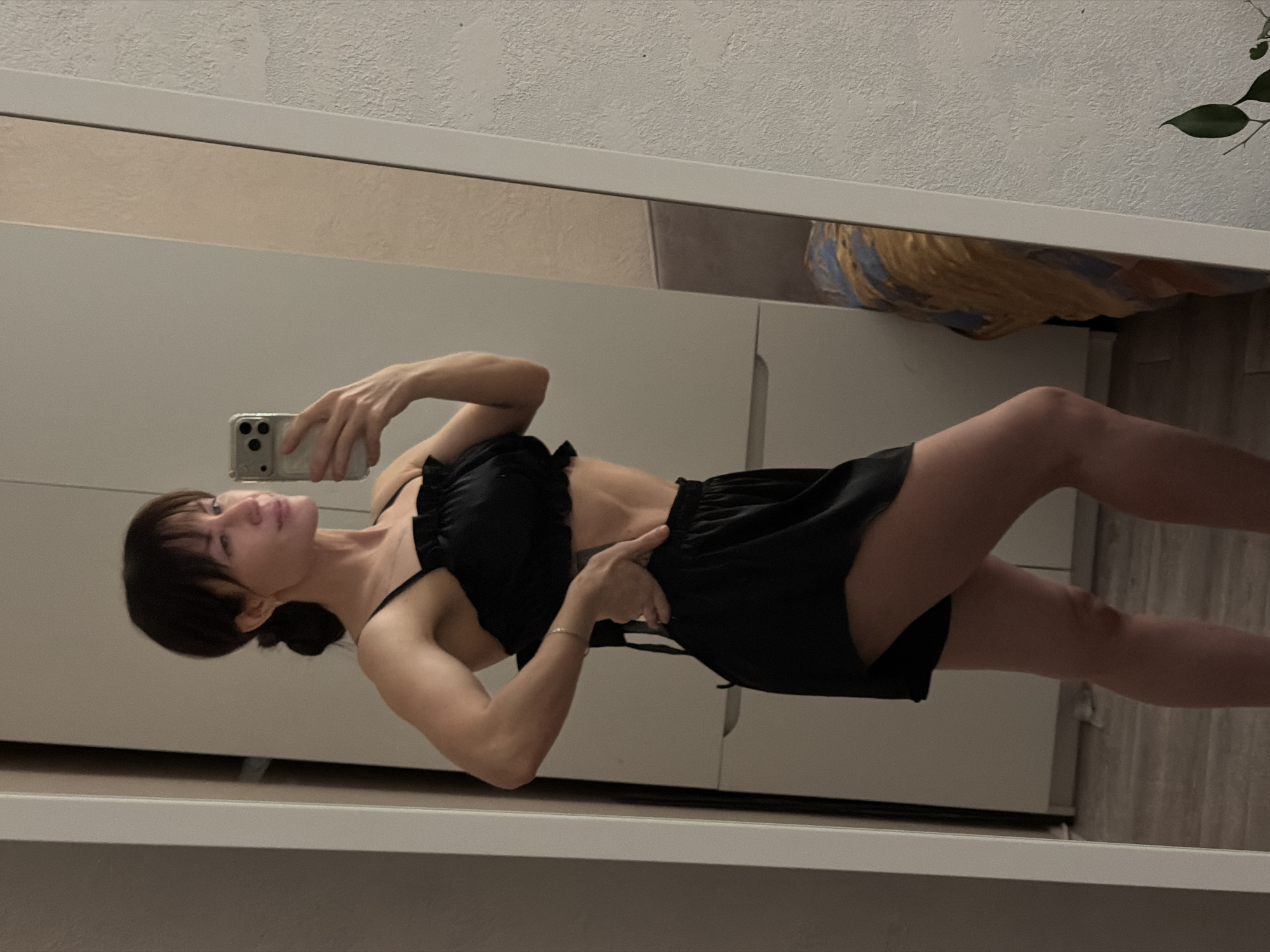 A woman is taking a selfie in a mirror. She is wearing a black two-piece outfit. The background includes a white closet or wardrobe and a hint of a plant. The location is not definitively discernible as Perm, Russia, from the visual information provided.