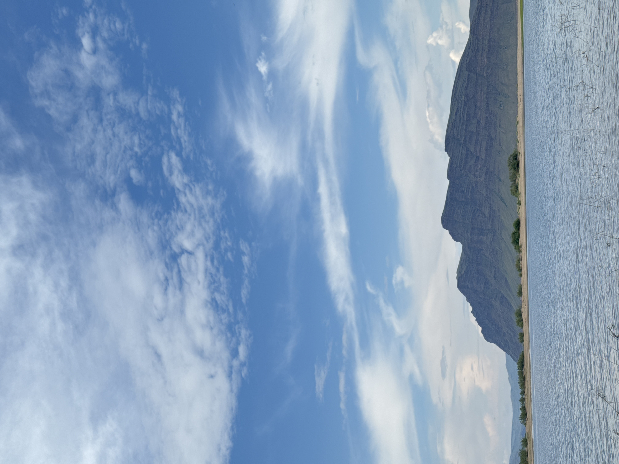The image depicts a serene, natural landscape under a vast sky dotted with white, wispy clouds. In the foreground, a calm body of water stretches across the frame, its surface rippled by a gentle breeze. The water appears shallow near the edges, with sparse reeds or dry vegetation emerging from it.
Beyond the water, a strip of sandy or light-colored land serves as a shoreline, sparsely dotted with small, green shrubs and trees. This vegetation gradually gives way to the imposing presence of a large, dark mountain range that dominates the background. The mountains are characterized by their rugged, layered slopes, with visible striations suggesting sedimentary rock formations. The peaks are jagged and uneven, silhouetted against the bright sky. The overall impression is one of natural beauty and tranquility, characteristic of a rural or wilderness setting. The location is identified as Nikolo-Petrovka, Russia.