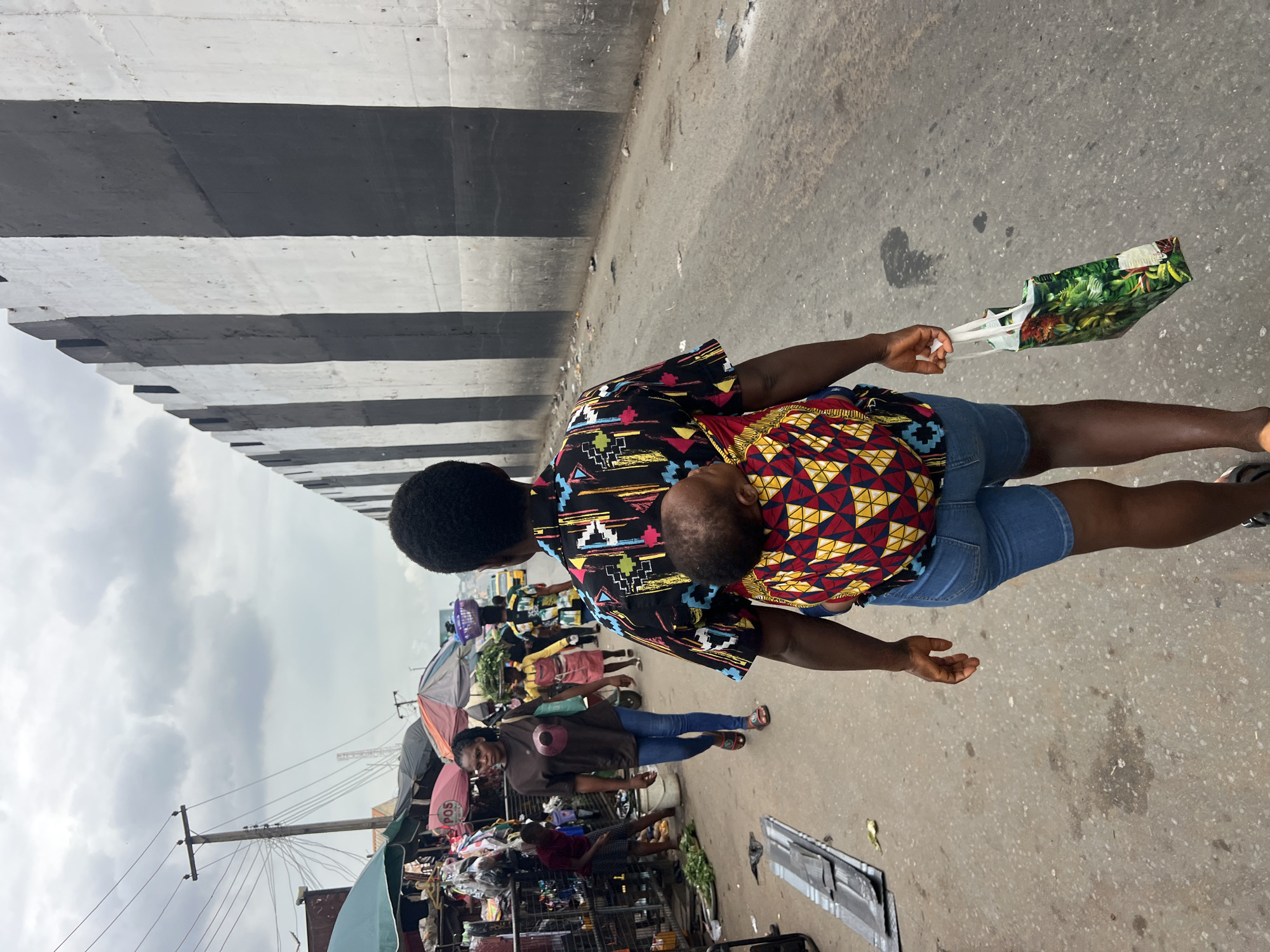 This image captures a vibrant street scene in Nkpor, Nigeria, likely during daytime under an overcast sky. The primary subject is an individual with dark skin, wearing a black shirt adorned with colorful abstract patterns and denim shorts. They are carrying a baby or toddler on their back, secured with a patterned fabric wrapper featuring red, yellow, and blue geometric designs. The child's head rests on the carrier's shoulder. In their right hand, the person holds a green patterned shopping bag with white handles. Their left hand is slightly open.
They are walking on a worn concrete or asphalt road, which shows some stains and debris. To the left, a bustling market unfolds, populated by other people, stalls, and umbrellas providing shade. One person in the mid-ground, wearing a brown top and blue jeans, looks towards the camera. Goods are displayed on various stands and racks, indicative of commercial activity. A pink umbrella visible in the background has the text "POS" printed on it. A wooden utility pole with numerous wires extends upwards against the sky, suggesting an active infrastructure.
Dominating the upper portion of the frame is a large concrete structure, presumably an overpass or bridge, characterized by bold horizontal black and white stripes. The sky above is cloudy, contributing to a muted light. The overall scene depicts a slice of daily life in a busy urban or semi-urban environment, with people engaged in commerce and movement.