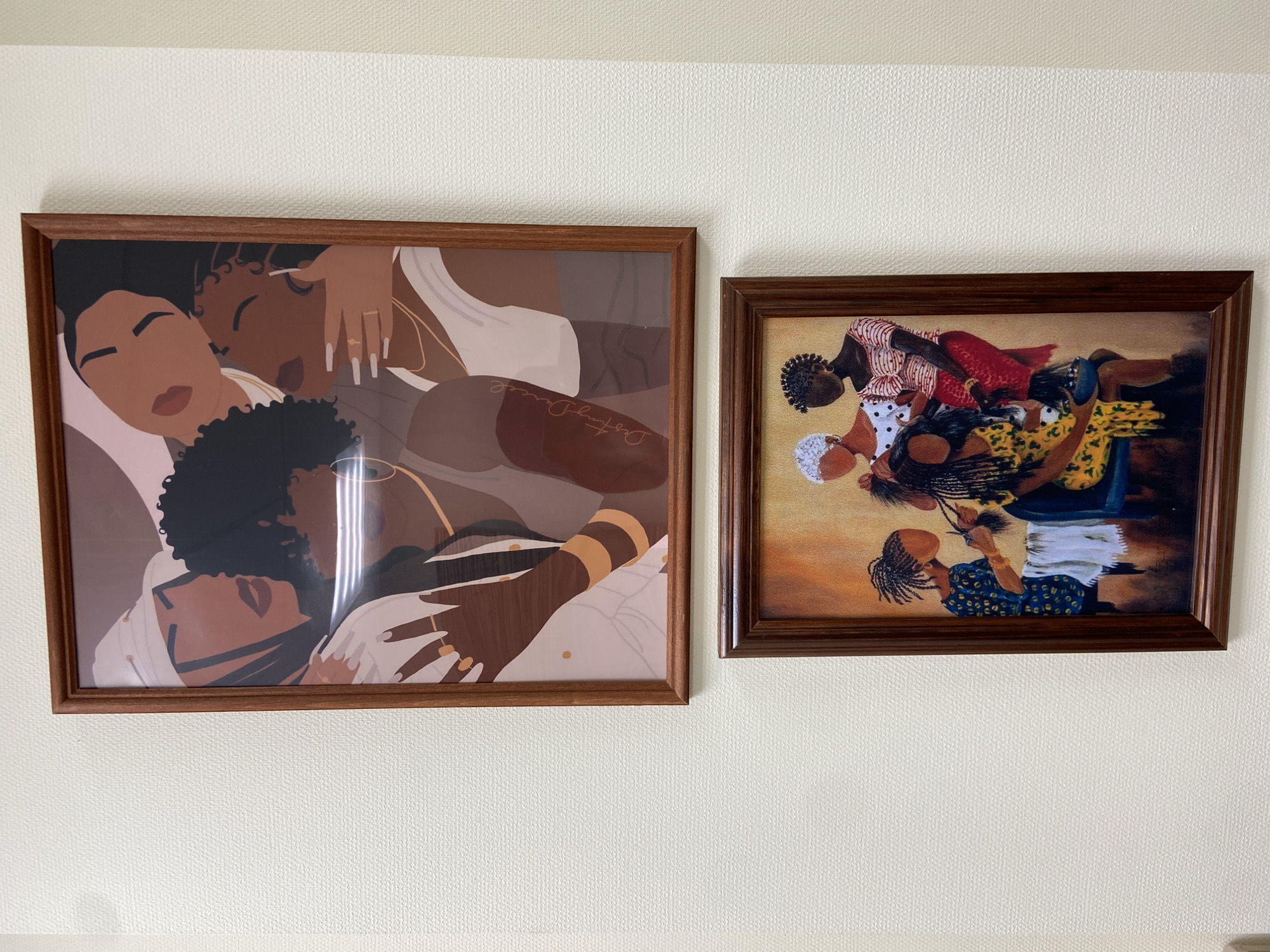 The image shows two framed artworks hanging on a white wall. The left frame depicts a stylized drawing of two dark-skinned women in a close embrace. The artwork features varied shades of brown, white, and gold, and one signature "DestinyDaniel". The right frame contains a painting of several Black women, possibly braiding hair, with vibrant clothing. This scene incorporates darker color shades with blue, red, yellow, and brown present. The frames on both artworks are made of dark brown wood. No location information is visible.