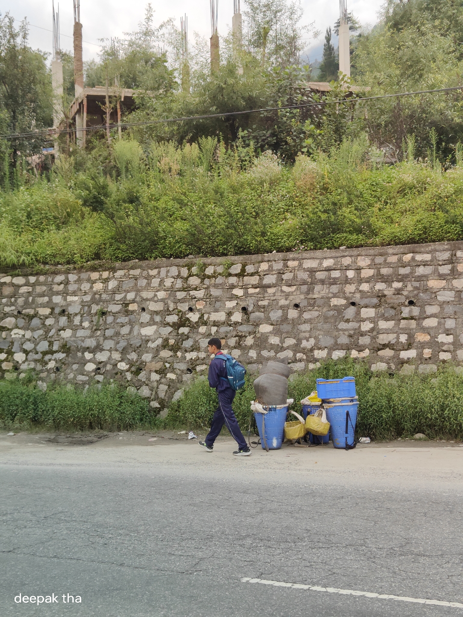 A young person in a dark blue tracksuit with a blue backpack is walking to the right on a paved road beside a stone wall. The person is positioned in the lower-middle portion of the frame. To the right of the person, there are several blue plastic containers stacked vertically, with yellow bags resting on them. A large gray pot is also stacked on one of the blue containers. Green vegetation covers the area behind the stone wall and also lines the edge of the road. In the background, there are trees and the unfinished concrete structure of a building with rebar protruding from the top. The sky is overcast. The location is identified as Kalath, India, based on the watermark in the lower-left corner.