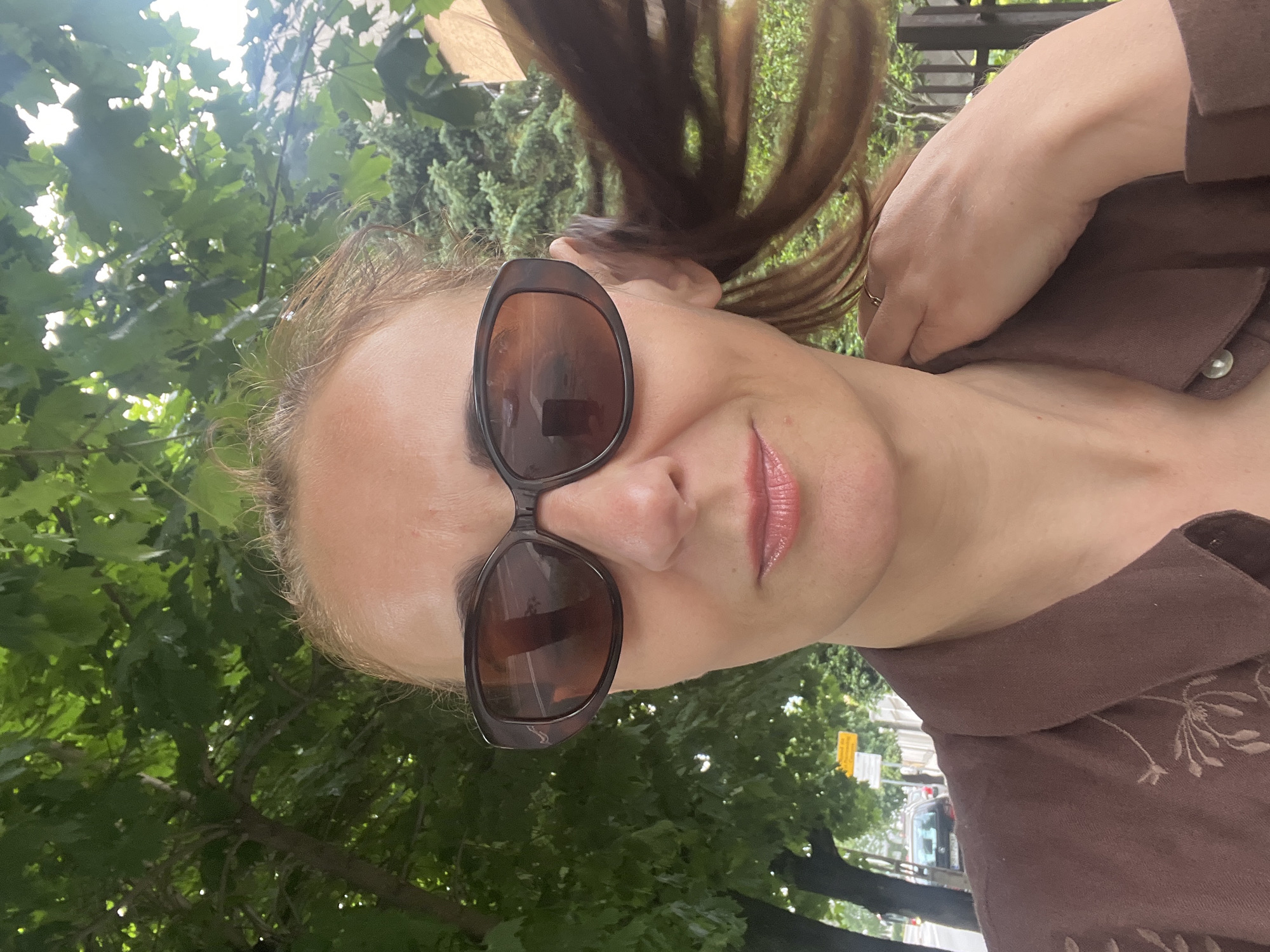 A woman with blonde hair pulled back and sunglasses is taking a selfie outdoors. She is wearing a brown collared shirt with embroidered details. Her right hand is touching her neck. Behind her, there are trees with green leaves, a street with cars, and a fence. The lighting suggests it is daytime.