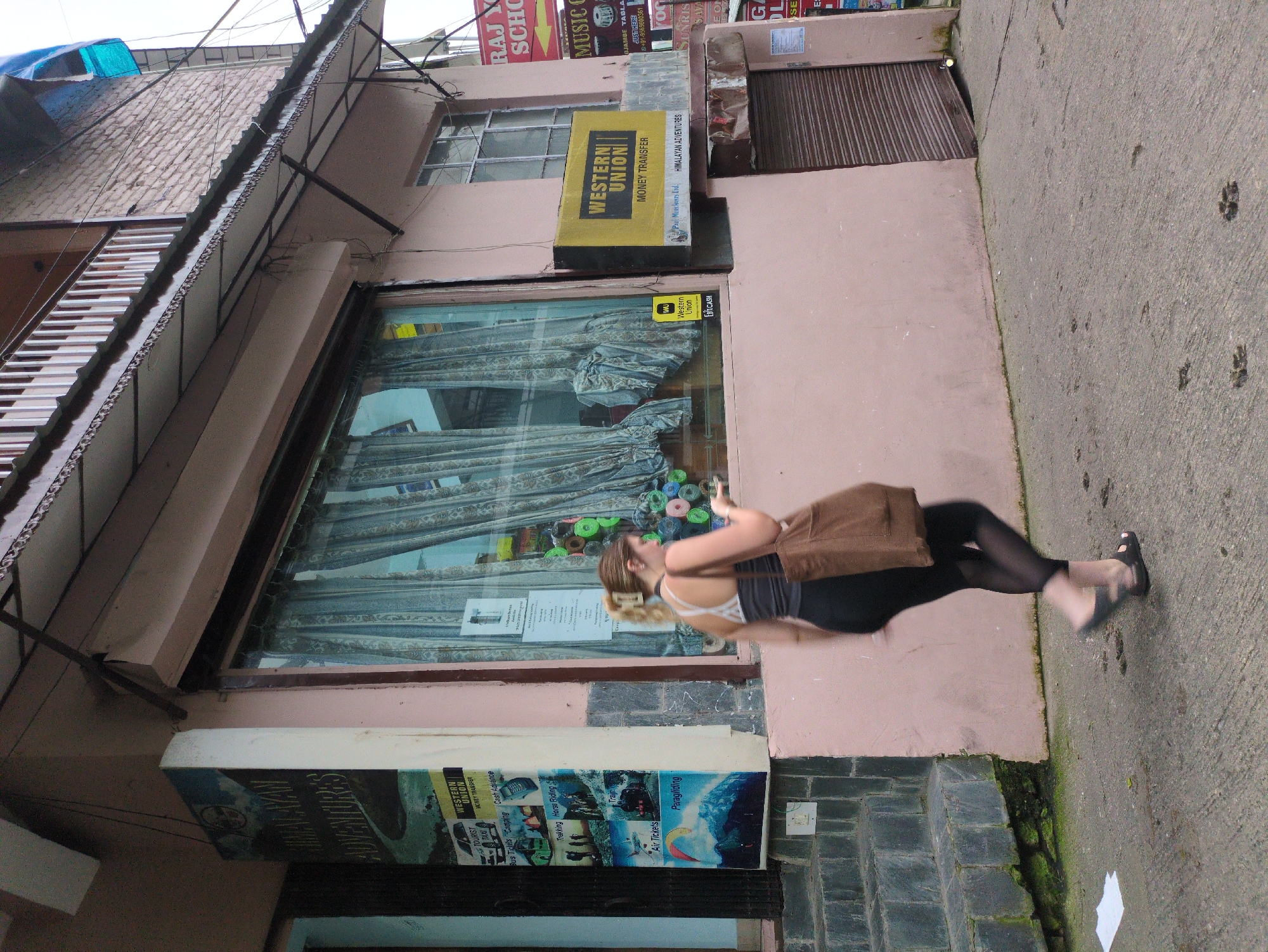 In Bhagsu, India, a woman walks from right to left in front of a pink building. She has blonde hair and is wearing a dark tank top, black leggings, and dark sandals. She carries a brown shoulder bag. The building features a large storefront window with blue curtains and displays various advertisements and signs. One prominent yellow sign reads "WESTERN UNION MONEY TRANSFER." To the left of the storefront, a vertical advertisement for "HIMALAYAN ADVENTURES" is visible, showcasing images of activities like paragliding and trekking. A set of stone steps leads up to a dark doorway on the far left. The ground in front of the building is a concrete sidewalk with visible paw prints. The overall scene suggests a commercial street in a town.