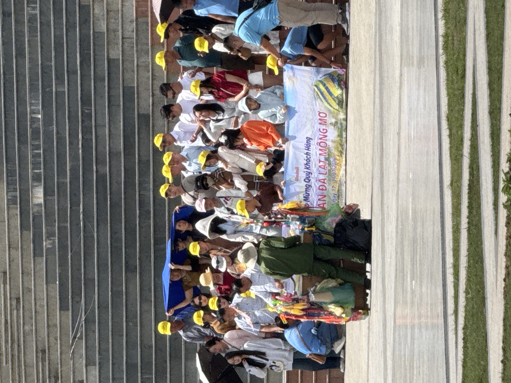 A group of approximately 30 people, most wearing yellow baseball caps, stand on wide stone steps under a bright sky. A large blue tarp is held aloft by several individuals in the center of the group. In front of the steps, a banner with Vietnamese text and an image of hot air balloons reads "MỪNG QUÝ KHÁCH HÀNG AN DA LAT MONG MO NGAY 17 - 19/07/2026". A table or cart is set up in front of the banner, displaying colorful items. Several people are gathered around this cart. To the left, a person in a blue shirt crouches down. To the right, a person in a green uniform stands near the cart, and further right, a person in a blue shirt and khaki pants interacts with someone kneeling. The scene appears to be a tourist gathering or event in Da Lat, Vietnam.