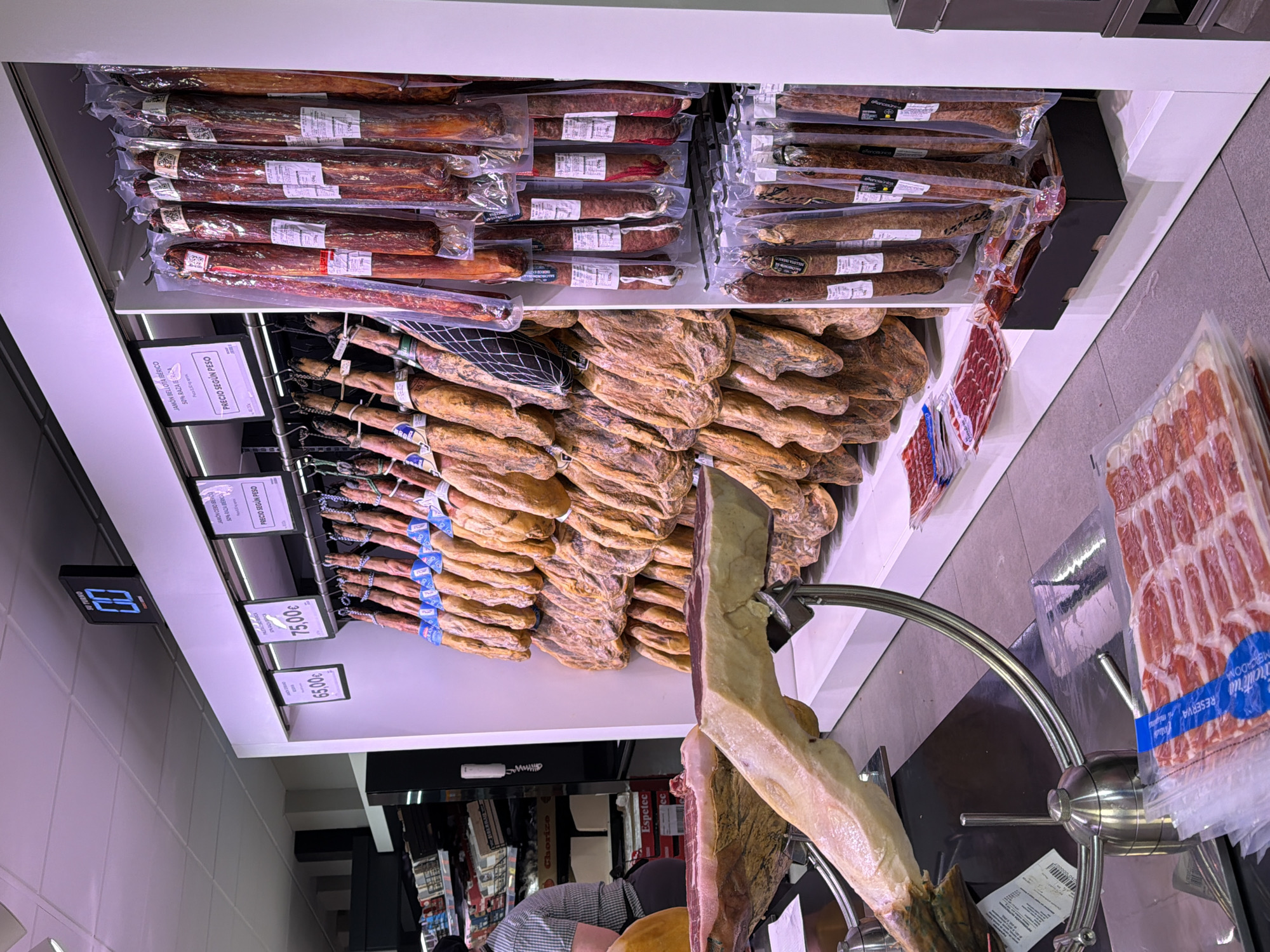 This image depicts a display of cured meats, specifically ham and chorizo, at a shop, likely a deli or supermarket. The hams are arranged on shelves, with some whole legs mounted on stands. The chorizos are packaged in plastic wrap. Price signs indicating prices per kilo are visible. A man in an apron can be seen behind the display. The OCR information suggests the meats are Iberian hams and chorizos, including "Jamón Bellota Ibérico" and "Jamón Cebo Ibérico." Further, the OCR information "SPA BAZA BERICA" is likely part of the brand. The location is in the city of Arroyo de la Miel-Benalmádena Costa, Spain.