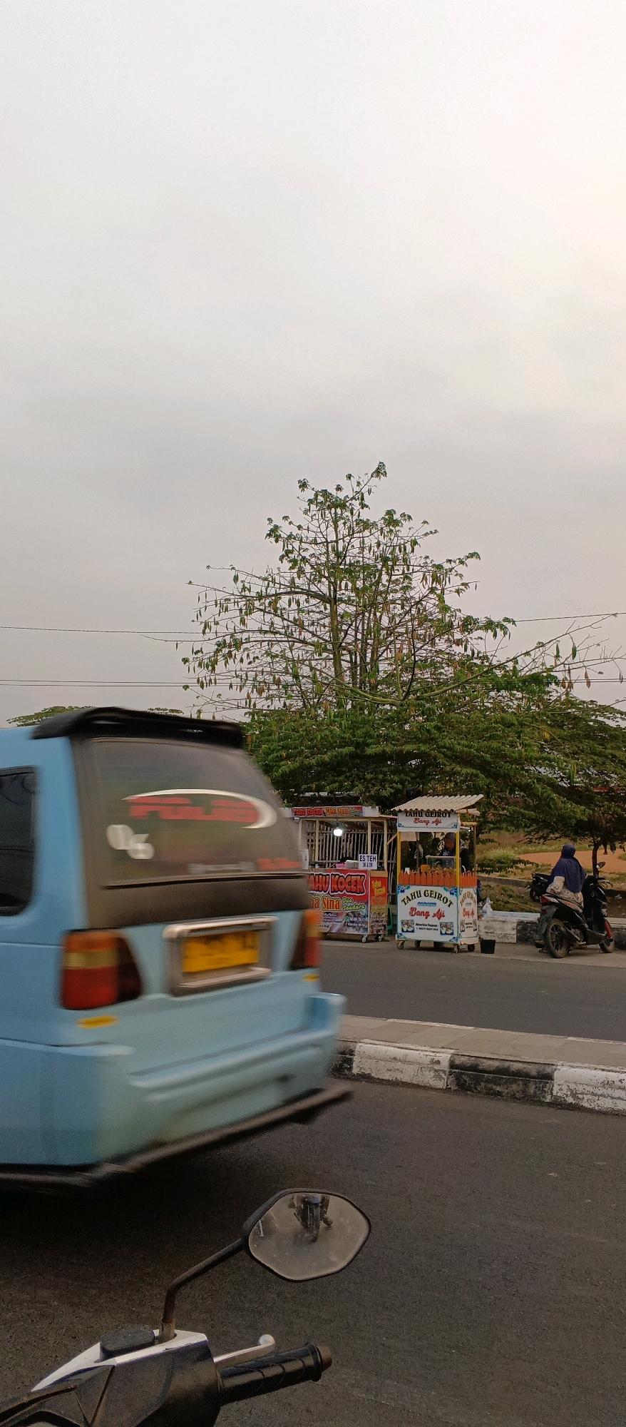 The image captures a street scene in Watubelah, Indonesia, under an overcast sky. In the foreground, the handlebars and a rearview mirror of a motorcycle are visible, reflecting other vehicles and the surrounding environment. A light blue van is driving past in the mid-ground, its motion blurred due to the camera's perspective and speed. Behind the van, a tree with prominent seed pods stands tall. To the right of the tree, two food carts are set up on the side of the road. One cart displays "TAHU KOLEK" and "ES TEH," while the other advertises "TAHU GEIROT BANG AJI." A person is seated on a motorcycle parked near the "TAHU GEIROT" cart. The street has a concrete curb and asphalt pavement, with power lines visible overhead. The overall impression is that of a typical roadside scene in a populated area of Indonesia.