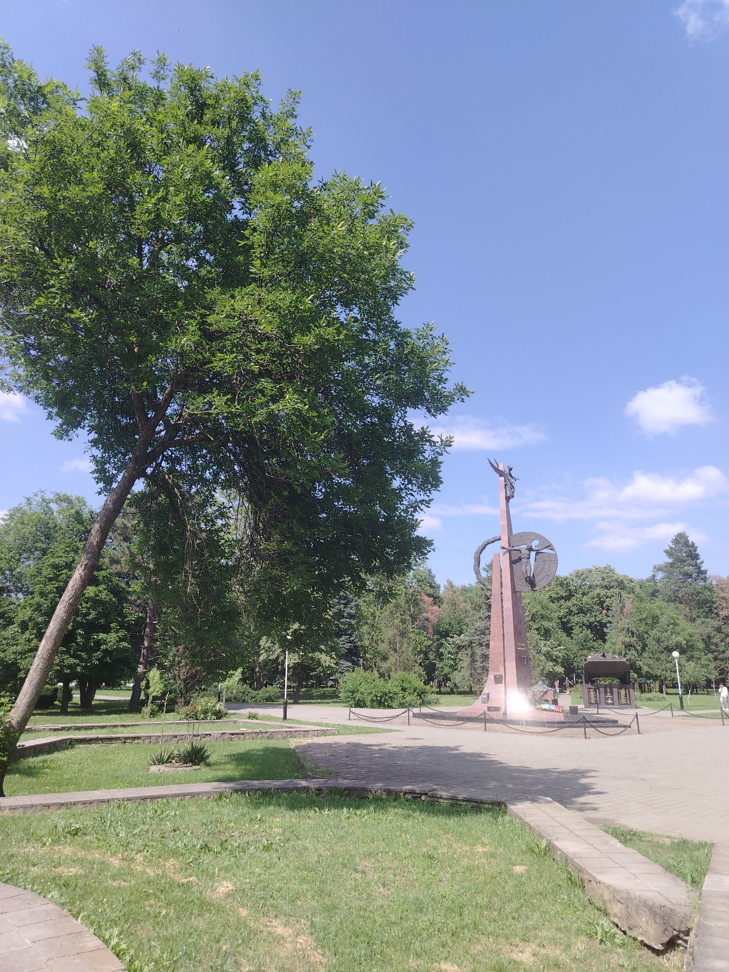 This is a medium shot photograph taken outdoors on a sunny day with a clear blue sky and a few scattered white clouds. The scene appears to be a park or public square, likely in Krasnodar, Russia.
The main subjects in the image are a large, leafy green tree dominating the left side of the frame, leaning dramatically towards the right. In the background, a tall, reddish-brown monument stands prominently. The monument is abstract in design, featuring geometric shapes and a circular element with a figure or symbol within it, topped by what appears to be a winged figure. To the right of the monument, there is a smaller structure with inscribed panels, possibly a memorial wall, and beyond that, more trees and distant buildings.
In the foreground, paved walkways and areas of green grass are visible. The grass has patches of brown, suggesting it might be a warm season. The overall setting is tranquil and park-like.
There are no people clearly visible engaged in any significant activity, though a small figure can be seen walking in the far distance on the right. The lighting suggests it is daytime, likely midday given the brightness and shadows. The atmosphere is serene and peaceful. No visible text is discernible in the image.