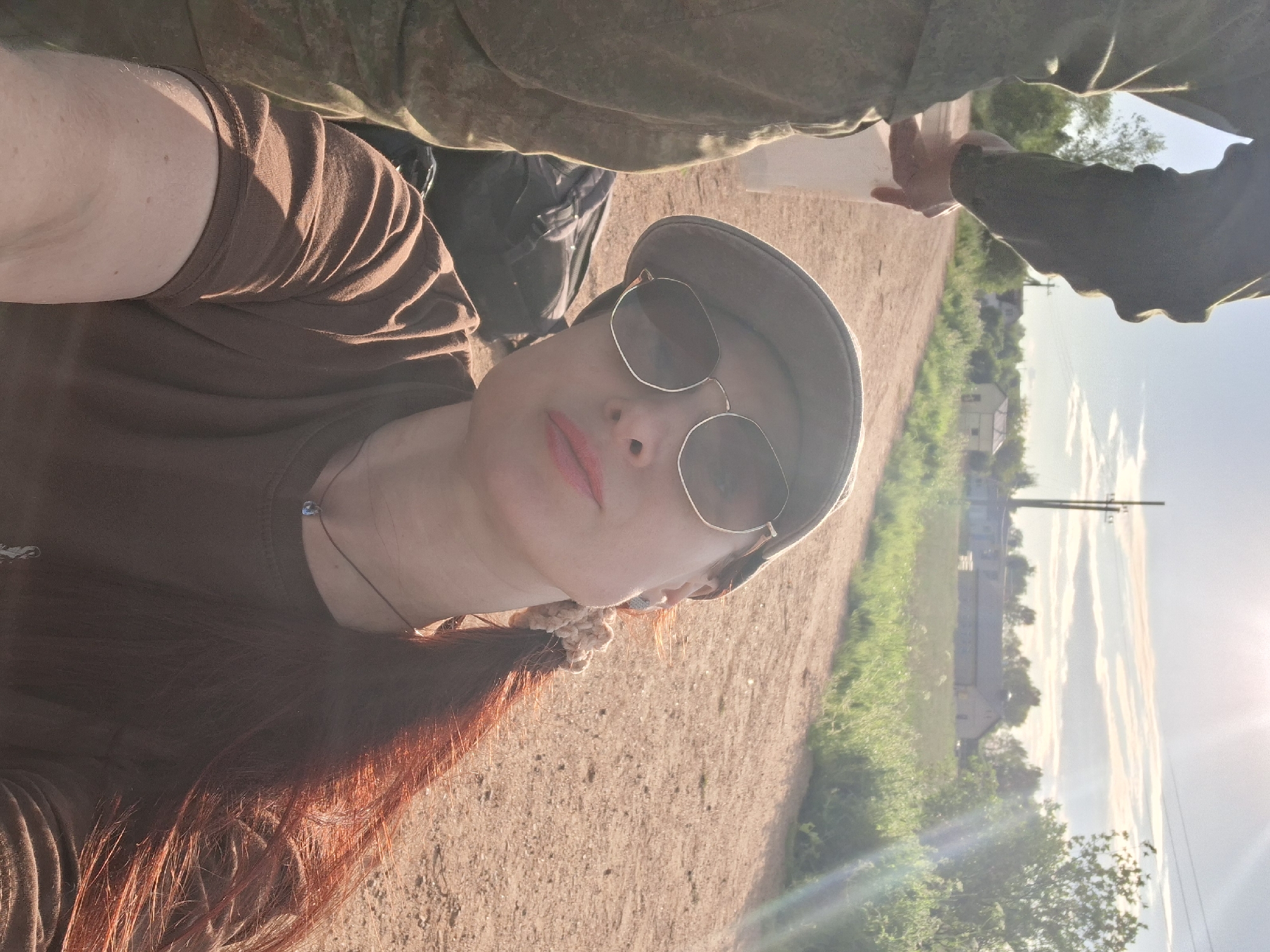 The image is a vertical selfie taken by a woman outdoors in a rural or semi-rural setting in Забалоцкі сельскі Савет, Belarus.
The primary subject is a woman with long, reddish-brown hair, wearing a brown t-shirt, a dark baseball cap, and round-framed sunglasses. She has red lipstick and a delicate necklace with a small blue bead. Her arm and shoulder are visible in the foreground, illuminated by warm sunlight.
Above and behind her, a second person is partially visible, wearing military-style camouflage clothing. This individual's arm and hand are extended, holding what appears to be a map or a piece of paper, suggesting they might be reviewing directions or information about the area. A backpack is also visible near this person.
The setting is an unpaved dirt path or road. To the right of the path, there is a vibrant green landscape with tall grass and trees. In the background, under a bright sky with some faint clouds, several distant buildings, likely houses, and a utility pole can be seen.
The time of day appears to be late afternoon or early evening. The sun is low in the sky, creating warm, golden light and casting long shadows. A prominent lens flare from the low sun is visible in the upper right portion of the image. The overall mood is bright and peaceful, suggesting travel or an outdoor activity. There is no visible text in the image.