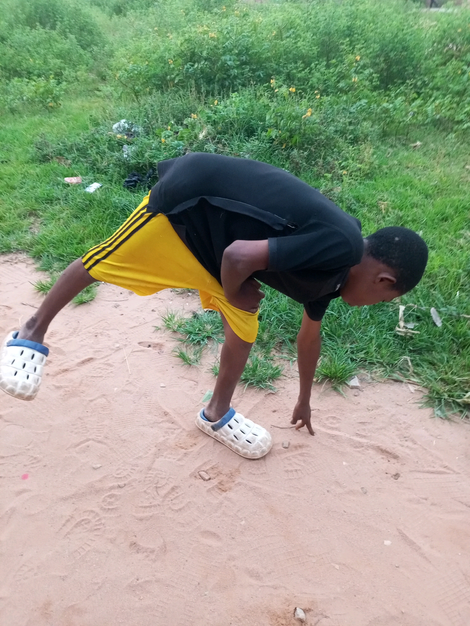 A young boy, dressed in a black t-shirt and yellow shorts with black stripes, is bending down on sandy ground. He wears white clogs with blue trim. His left leg is extended behind him, and his right hand is reaching towards the ground. The setting appears to be outdoors with a grassy area in the background, which includes some patches of yellow flowers and scattered debris. The sandy ground has numerous footprints imprinted on it. The image suggests a casual, possibly playful, moment, with the boy focused on something on the ground. The lighting indicates it is daytime, and the weather appears to be clear. The location context is Tempure, Nigeria.