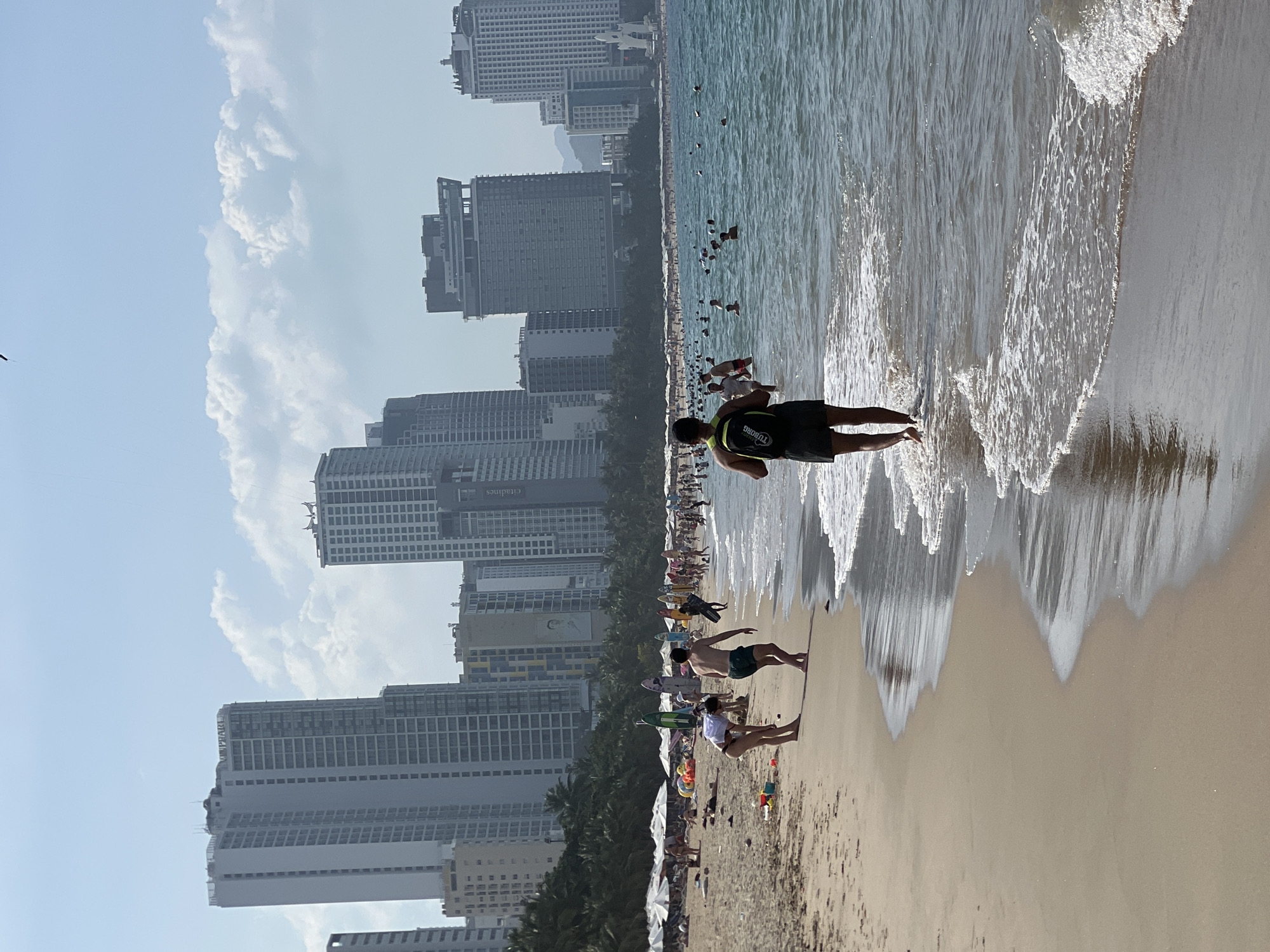 The image depicts a vibrant daytime beach scene in Nha Trang, Vietnam, identified by the "VINPEARL" branding on one of the prominent high-rise buildings in the background.
The sky is a bright blue with scattered white cumulus clouds. The horizon is dominated by a dense cityscape featuring numerous tall, modern buildings, including residential towers and hotels. A strip of green palm trees separates the buildings from the sandy beach. One large building in the mid-distance displays a billboard with a person's image. Distant mountains are faintly visible on the far right.
The wide, light-brown sandy beach is populated with numerous individuals. A section of the beach is equipped with multiple white beach umbrellas and lounge chairs. Various colored surfboards and paddleboards are upright in the sand. Small plastic beach toys are visible near the waterline.
In the mid-ground, a woman in a white top and dark swimwear is bending over on the sand near the water. Beside her, a man in dark shorts walks barefoot towards the water. In the foreground, viewed from behind, a man wearing dark shorts and a black backpack (with "TU DONG" visible) is walking barefoot into the shallow, light blue-green ocean water. Gentle waves with white foam are breaking along the shoreline. Numerous people are present in the water, swimming and wading. The scene is brightly lit by direct sunlight.