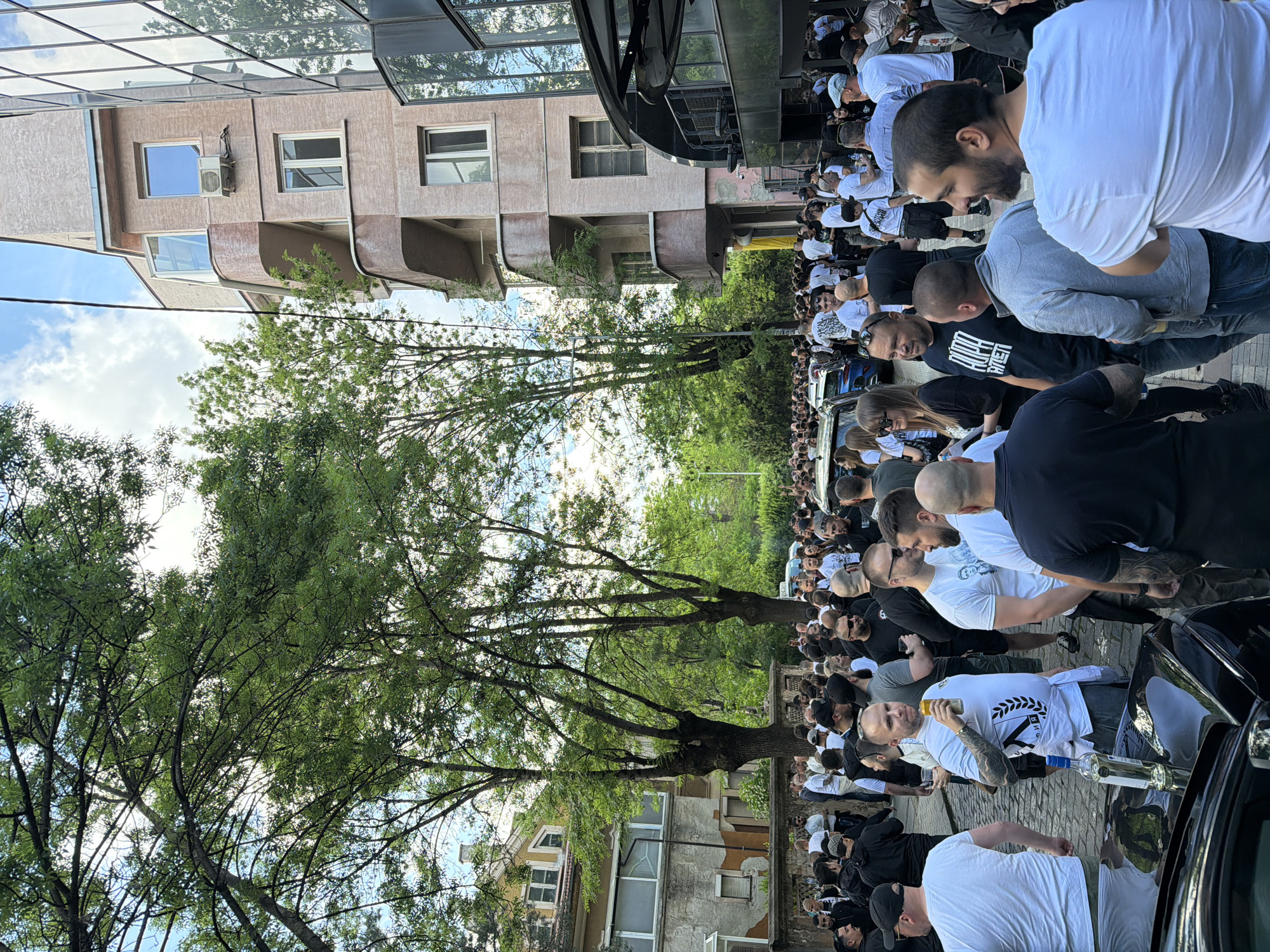 A large crowd of people, mostly men, fills a street bordered by buildings and trees. The crowd is largely dressed in black and white clothing. Cars are visible along the edge of the crowd. The location is Sofia, Bulgaria.