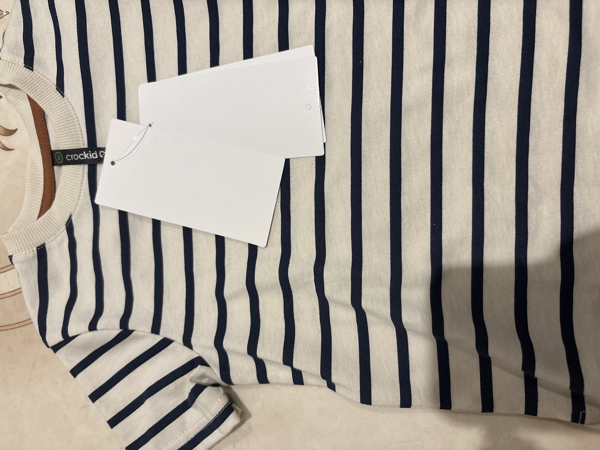 The image shows a striped shirt, primarily white with navy blue vertical stripes, lying on a surface. A tag with the brand "crockid" is attached near the neck of the shirt. Two rectangular white tags are also present, attached to the shirt. The background surface appears to be off-white or beige. The image does not provide sufficient information to confirm its location in Chaikovsky, Russia.
