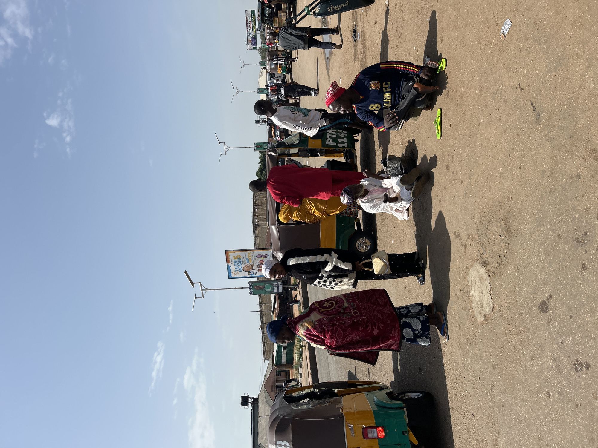 A street scene depicts a bustling outdoor area with several people and motorized rickshaws, commonly known as "Keke NAPEPs" or auto-rickshaws. The environment appears to be a marketplace or a transportation hub in a city. The ground is paved with a mix of asphalt and dirt, showing signs of wear and tear. Several parked and moving Keke NAPEPs are visible, painted in various colors like yellow, green, and brown.
In the foreground, on the right, two men are seated on the ground. One is wearing a blue and yellow long-sleeved shirt with "FC" visible on it, a red beanie, and black pants. He is looking to his left and has a pair of green flip-flops near him. The other man, to his left, is wearing a white robe and a patterned cap, with his legs crossed and holding a stick. A dark bag is beside him.
To the left of the seated men, a woman is standing and facing left, wearing a dark patterned outfit and a white headscarf, carrying a small beige purse. Next to her, a woman is wrapped in a patterned red blanket and wearing a blue headscarf.
Further back, other individuals are standing or walking. A man in a bright red traditional outfit is near a yellow and green Keke NAPEP, possibly interacting with someone inside. Another man in a white long-sleeved shirt and blue shorts stands near the same Keke NAPEP. A woman in a black and white patterned jacket and black pants is standing near another Keke NAPEP. In the background, more Keke NAPEPs and some buildings are visible, along with streetlights and billboards. One billboard prominently displays "TALENT UNCAGED 5 YEARS" and an image of a man. The sky is clear blue with a few scattered clouds, suggesting a bright, sunny day. The overall atmosphere is one of everyday activity and commerce.