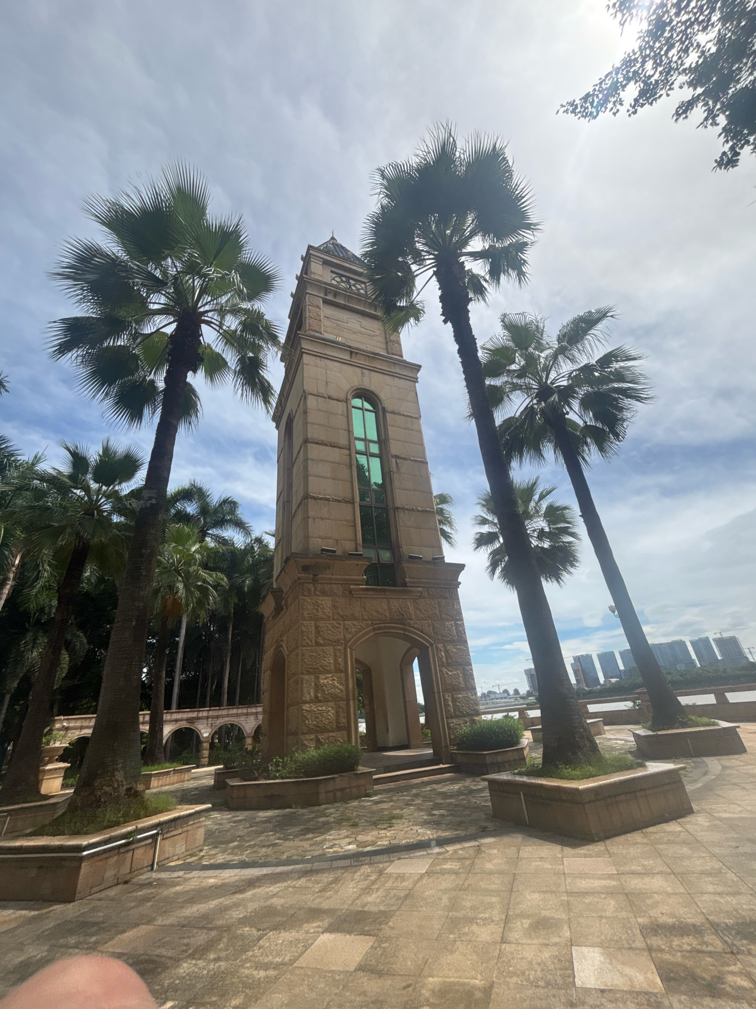 An eye-level, wide shot captures a tall, stone clock tower in Nanhai District, China, on a partly cloudy day. The clock tower, constructed of rough-hewn stone blocks, features a green, arched window near its top and an arched entryway at its base. Several tall palm trees with distinctive fronds are positioned around the tower, some in planters. The foreground is paved with large, square stone tiles. In the background, a body of water is visible, beyond which lies a modern cityscape with numerous high-rise buildings and construction cranes. The overall scene suggests a park or public space near a waterfront in an urban environment.