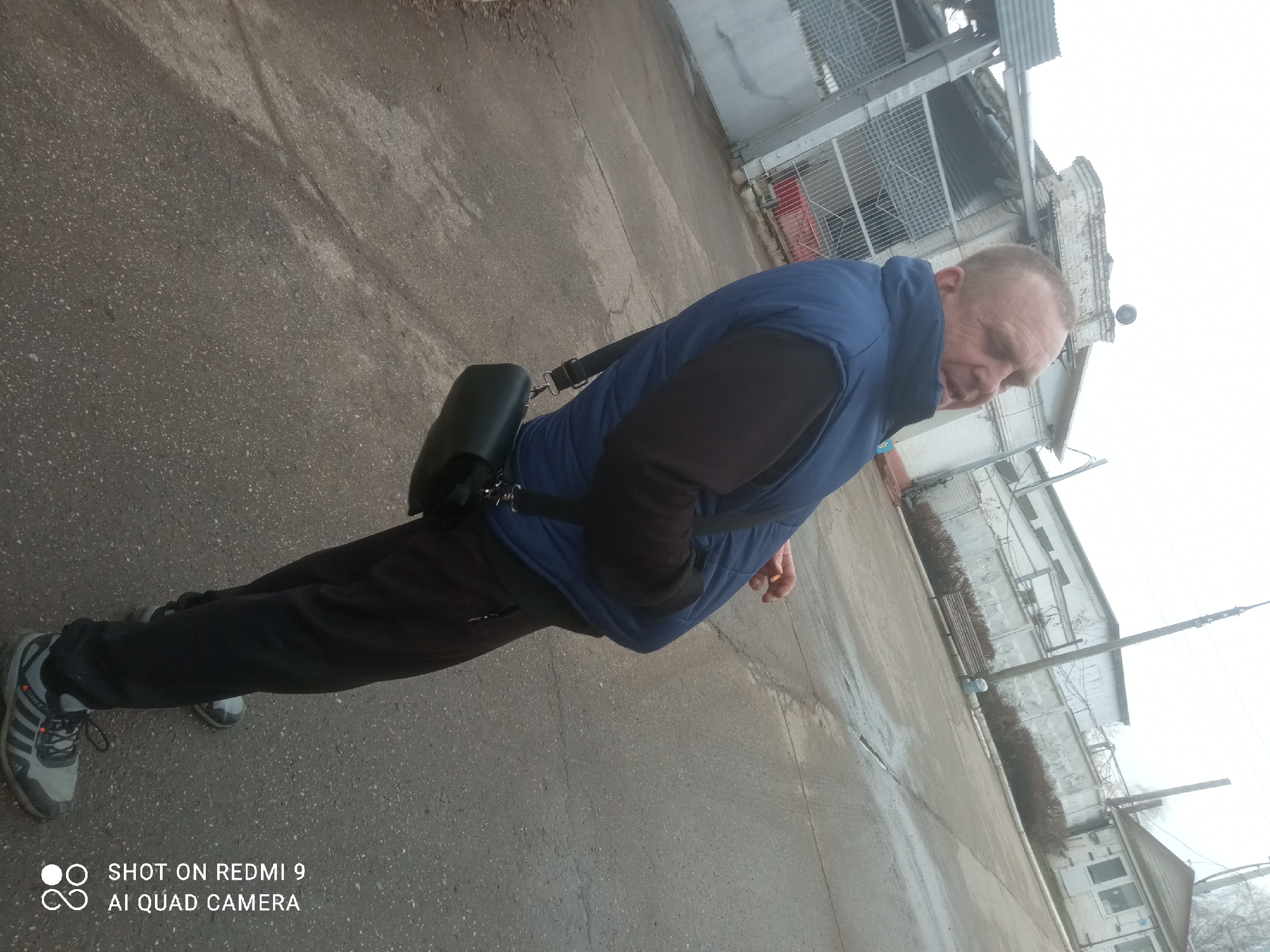 A middle-aged man stands on a concrete surface in an urban environment. He is wearing a blue vest over a black long-sleeved shirt, black pants, and grey athletic shoes with black stripes. A black bag hangs from a strap across his body. The man looks down.
Behind the man, there is a building with a metal cage-like structure and white walls. The area is paved and appears to be somewhat industrial or commercial.
Based on the camera tag, the image was taken on a Redmi 9 AI Quad Camera. The location is Chaikovsky, Russia, as confirmed in the metadata.