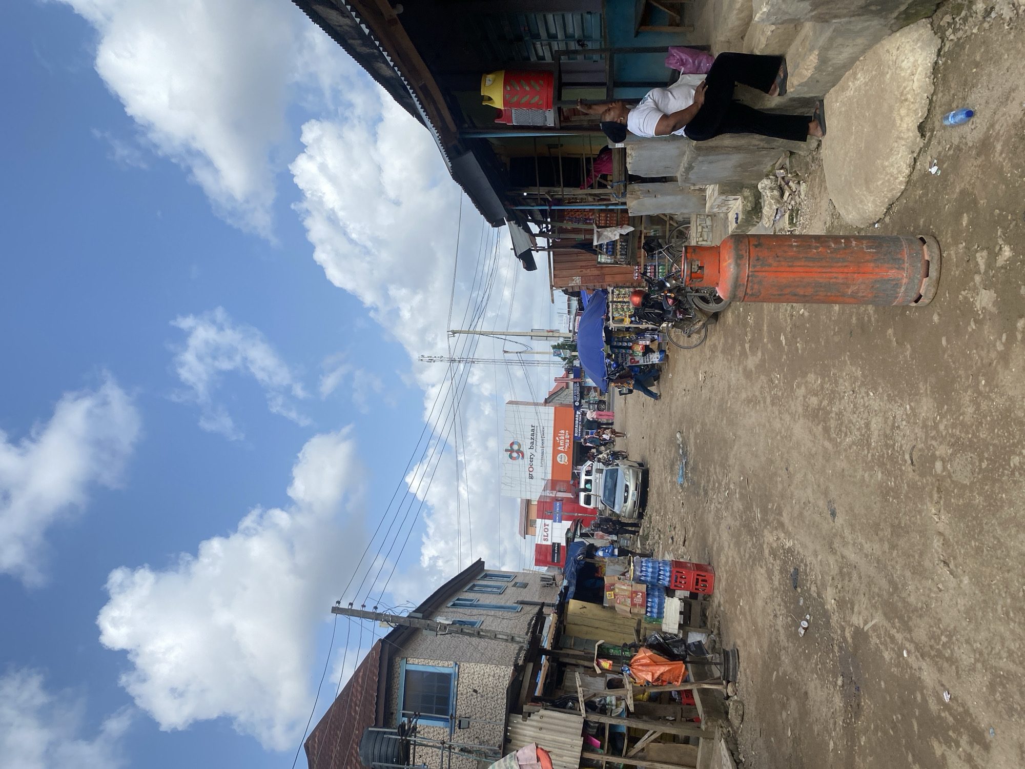 The image depicts a street scene in what appears to be a busy area in Lagos, Nigeria. The sky is bright blue with scattered clouds. Overhead power lines are visible. Buildings line both sides of a dusty dirt street. Various shops are open, selling goods such as groceries, beverages in crates, and more. A white van and several motorcycles navigate the street. People are seen walking and conducting business. An orange gas cylinder is leaning against a step where a man is lying down. Advertising signs are present on taller structures.