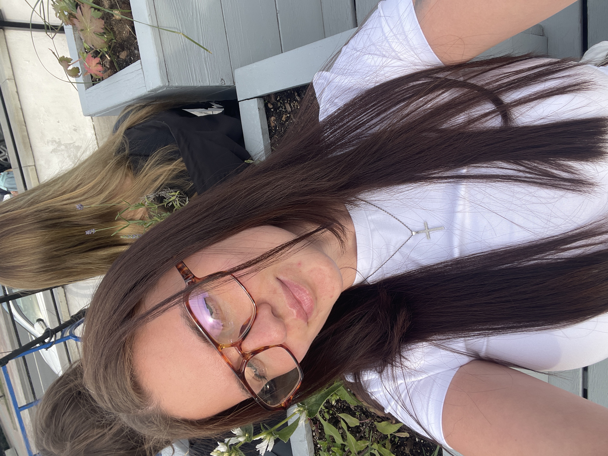 A woman with long, dark brown hair and glasses is taking a selfie. She is wearing a white shirt and a silver cross necklace. Her face is turned towards the camera. In the background, there are plants in a planter box and another person with blonde hair, but their face is not visible. The location appears to be outside in a garden or patio area. There is no evidence to suggest this image was taken in Moscow, Russia.