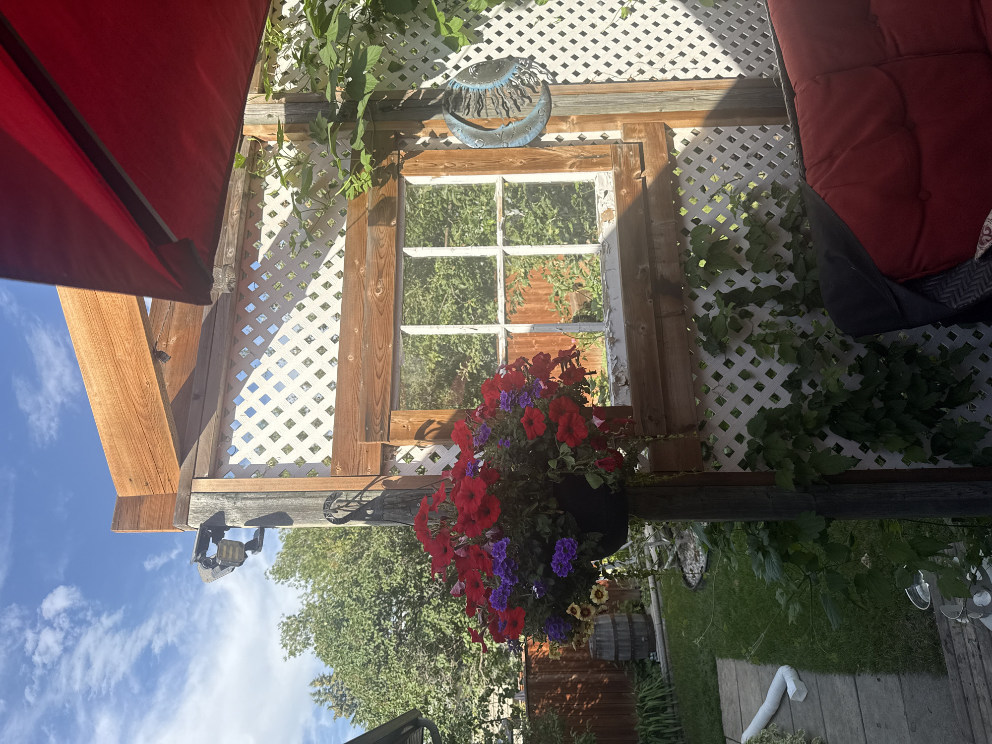 This is a photograph of an outdoor patio scene. In the foreground, a red cushioned outdoor sofa is partially visible. To the left, a wooden post supports a hanging basket filled with red and purple flowers. Behind the flowers, a white lattice fence with a wooden frame surrounds a section of the patio. A vintage window with peeling white paint is set into the lattice. To the left of the lattice, a section of lawn, a wooden fence, and a wine barrel are visible. Above the lattice, a wooden pergola structure extends towards the sky. A red umbrella is partially visible in the upper right corner. In the upper left quadrant of the image, a solar-powered motion sensor light is mounted on a wooden post. The scene is bathed in sunlight, suggesting a daytime setting. The photo was taken in Calgary, Canada.