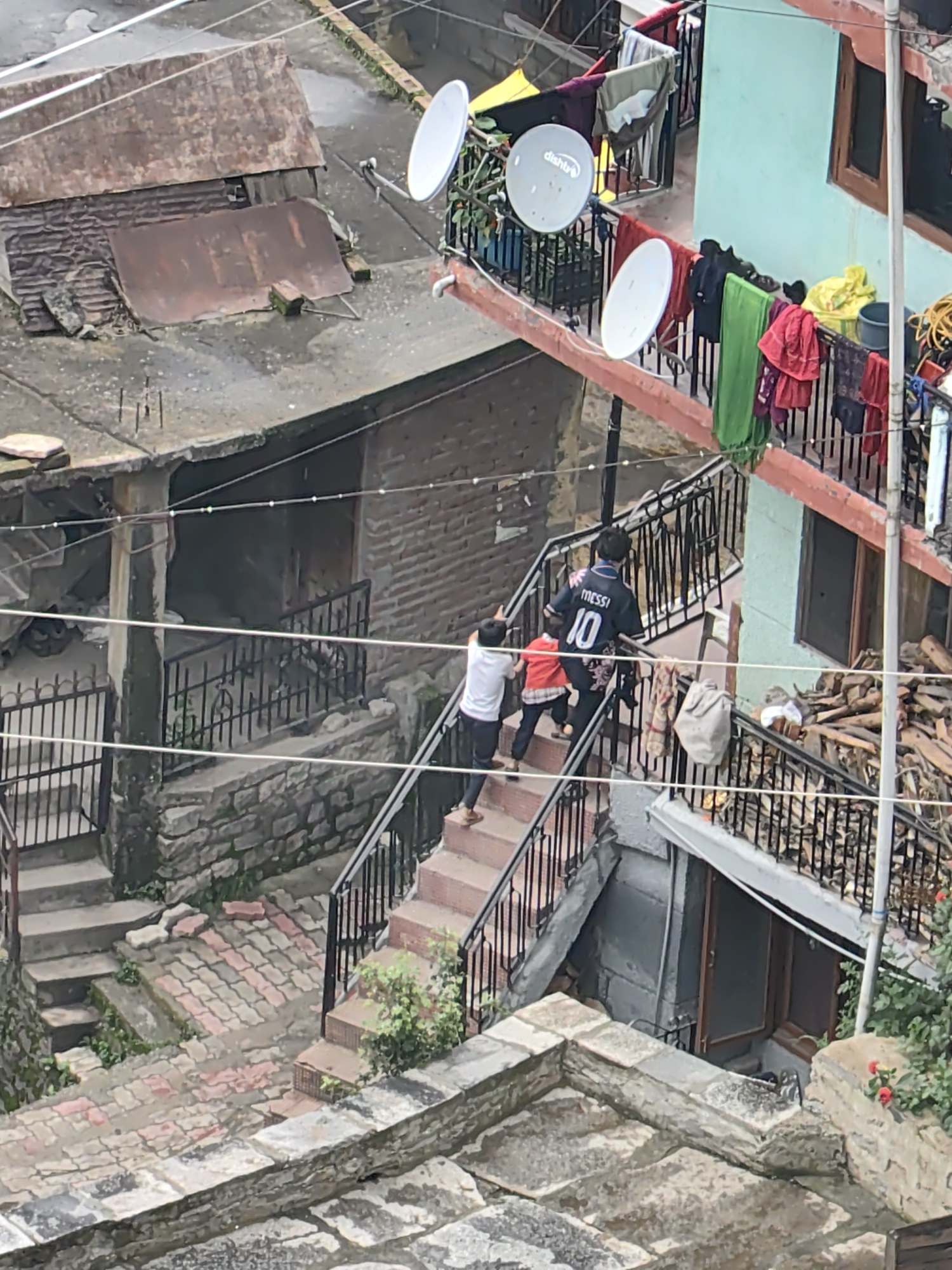 In Vashisht, India, a group of children is ascending a multi-level staircase constructed with stone blocks and red-tiled risers. The staircase is bordered by a black metal railing. A young boy in the lead wears a dark t-shirt with "MESSI 10" printed on the back. Following him is a child in a red jacket and black pants, and another child in a white t-shirt and black pants. The surrounding environment features multi-story buildings with weathered facades, some with corrugated metal roofs. Laundry hangs on balconies, and satellite dishes are visible. The ground level of the staircase shows a pathway paved with interlocking stones. The overall scene depicts a residential area with a sense of everyday life.