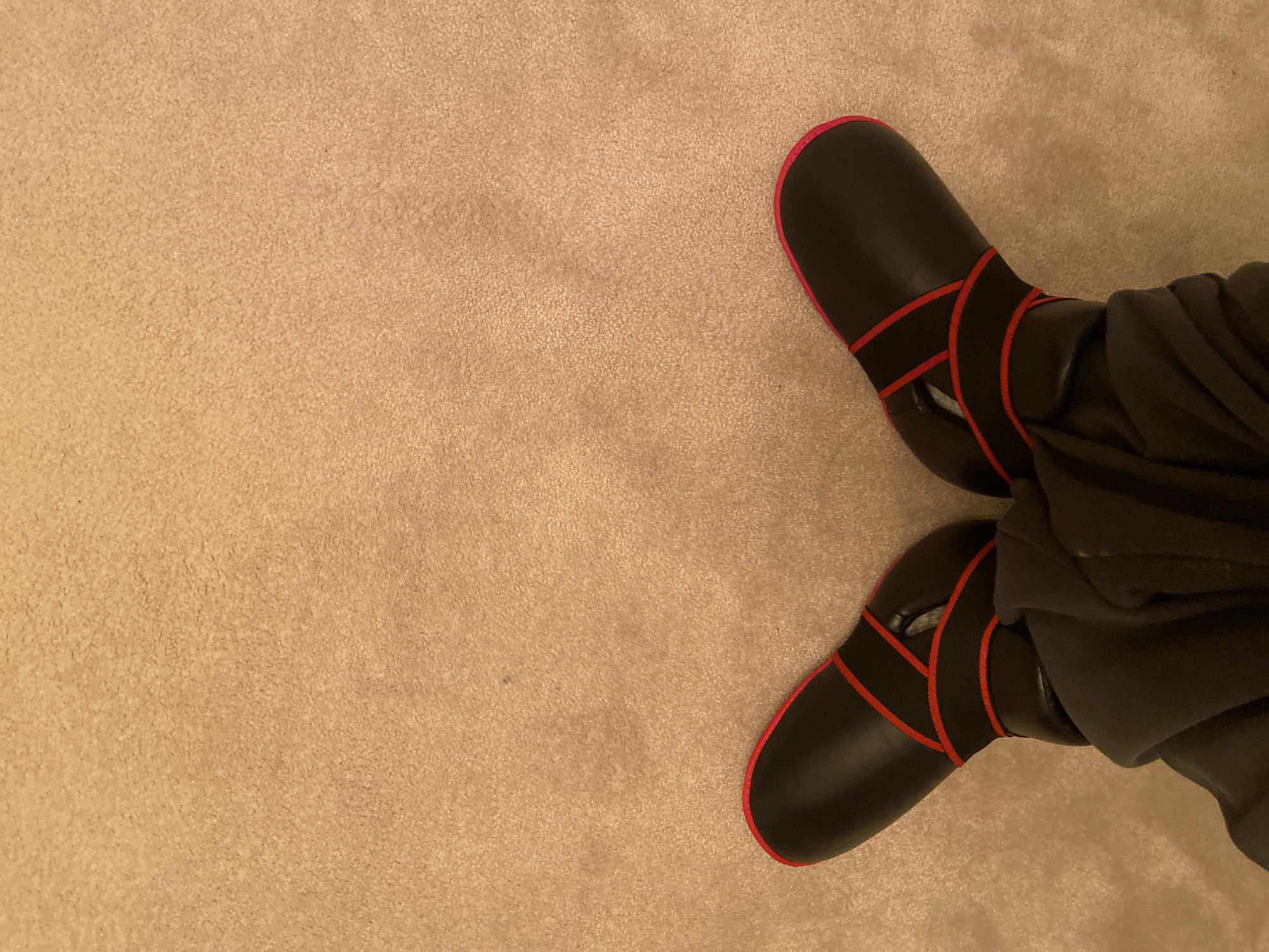 The image shows a first-person perspective of a person's feet resting on a beige carpet. The person is wearing black shoes with red trim and straps. They are also wearing dark-colored pants. The location cannot be determined from the image alone, and there is no evidence to indicate it is within Welwyn Hatfield, United Kingdom.