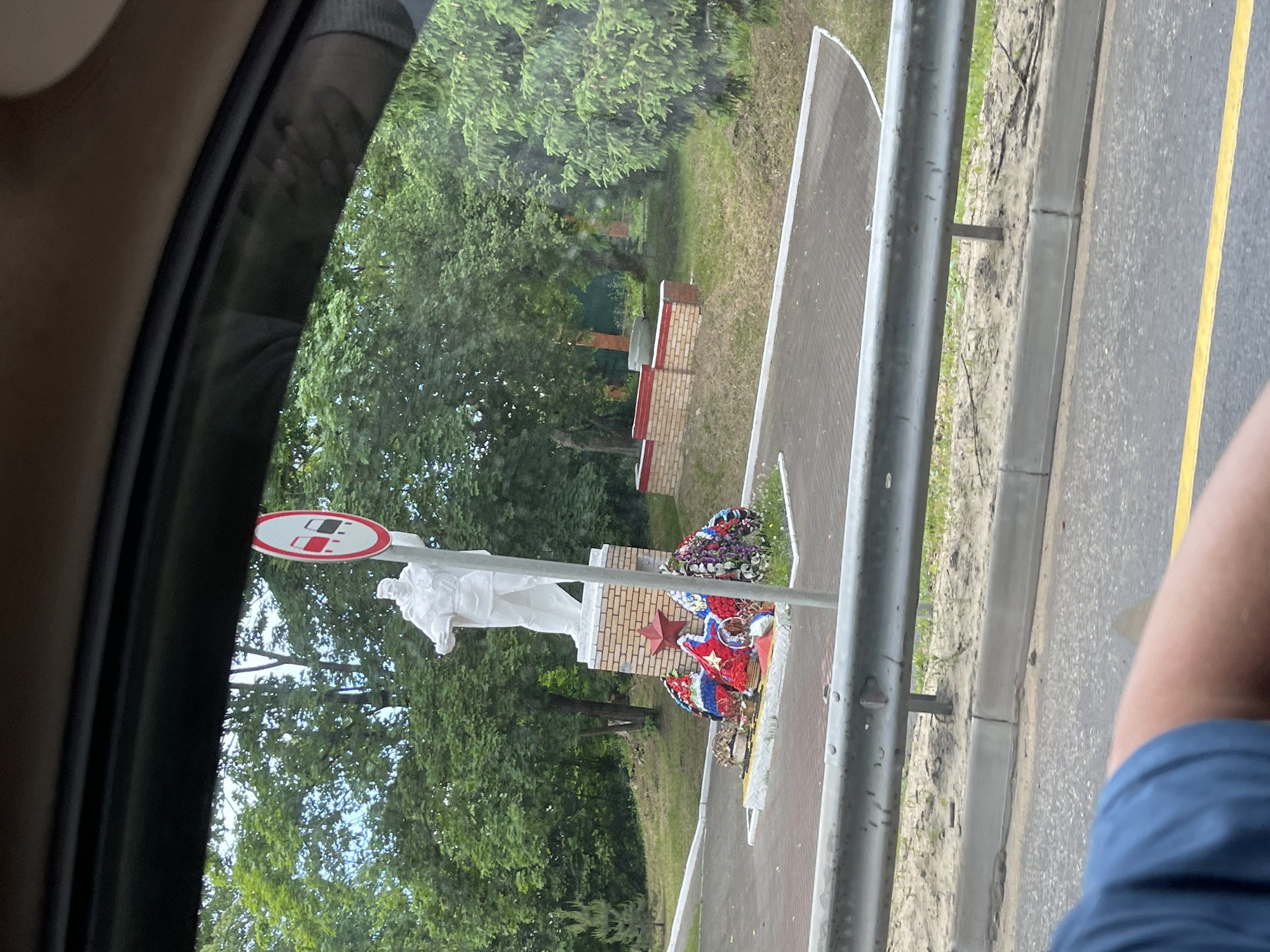 Captured from within a vehicle, the image reveals a monument commemorating soldiers in Semyonovskoye, Russia. The monument comprises a white statue of a soldier set on a brick pedestal. Wreaths adorn the base. A sign is visible above the statue. Trees form the backdrop. A road with a yellow line is seen along with a metal guardrail. An arm wearing a blue shirt is visible in the lower right corner of the frame. The scene is viewed through the car's window frame.