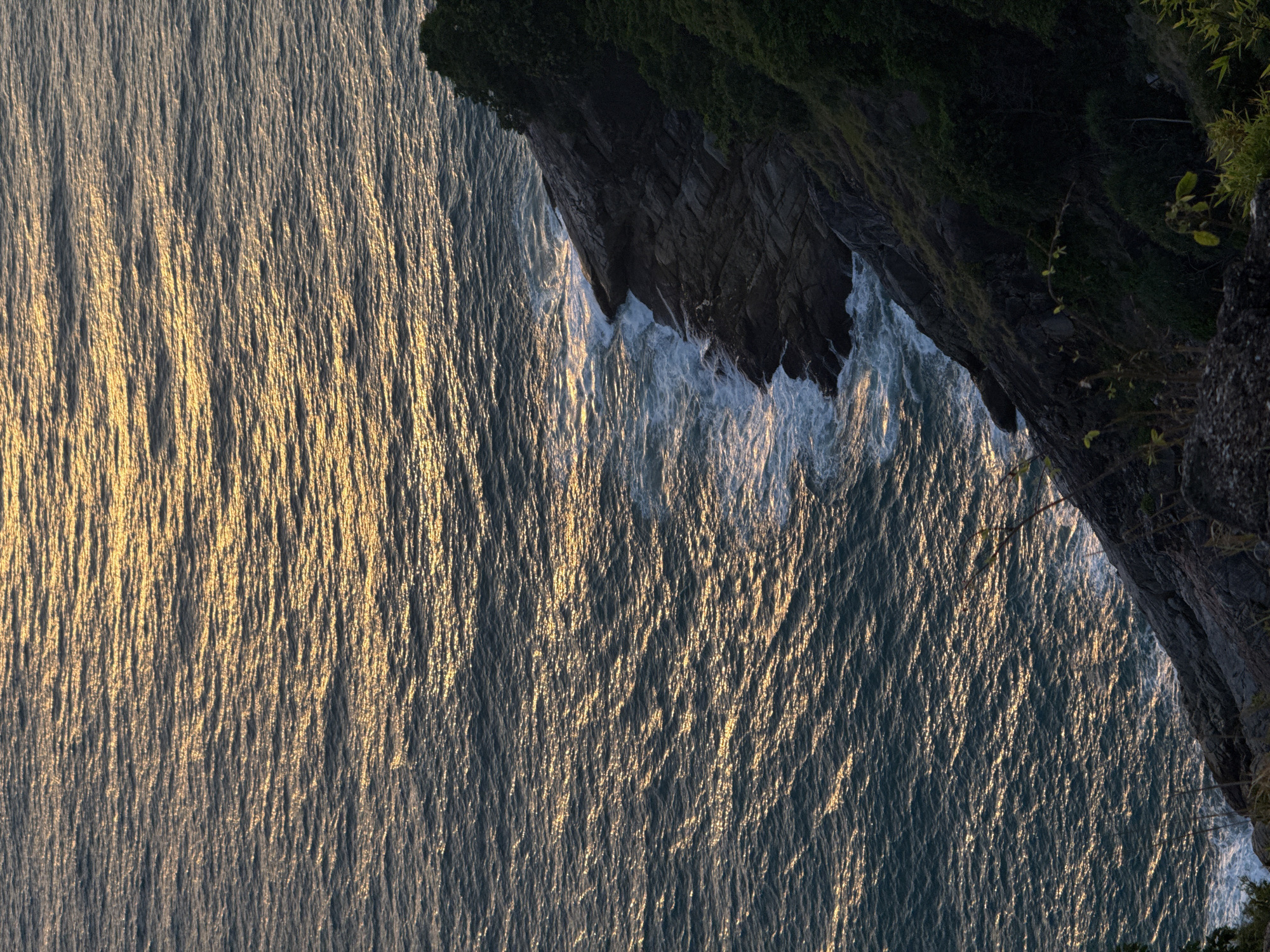 The image shows a view of the sea from a cliff overlooking Karon Beach in Karon, Thailand. The sun is setting, casting a golden light on the water, which has gentle waves. A rocky cliff covered in lush green vegetation slopes down to the sea. The scene is tranquil and natural.