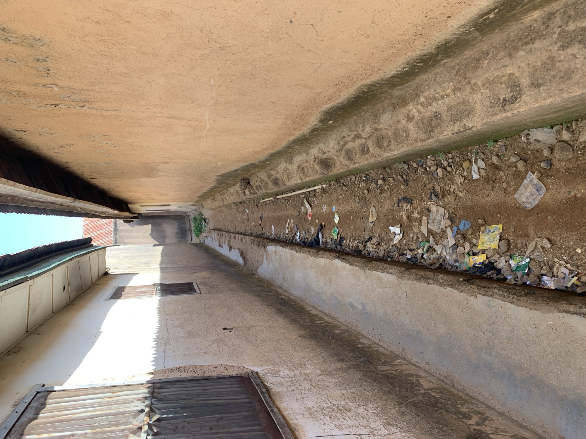 The image depicts a narrow alleyway in Rafin Par, Nigeria, between two buildings. One building is made of concrete and the other building has white walls. The alleyway has a dirt-filled trench along its length that is full of trash.