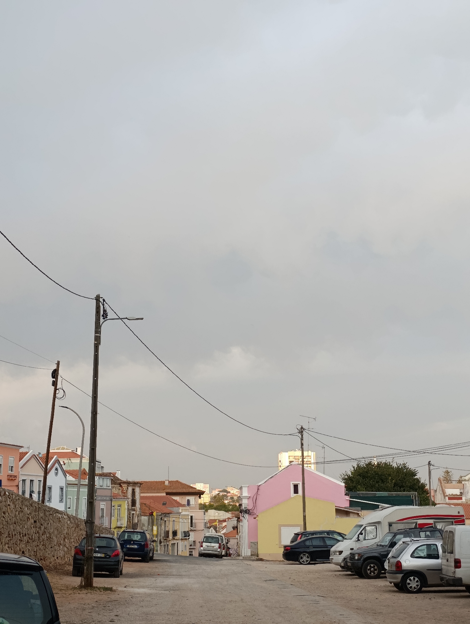 The image depicts a street scene in Setúbal, Portugal, characterized by a row of houses and parked vehicles. The sky is overcast with gray clouds, suggesting a potentially dull or rainy day. In the foreground, several cars are parked along the side of a dirt and gravel road. These include a black hatchback, a dark blue SUV, a silver compact car, and a white van. Further down the street, more vehicles are visible, including what appears to be a camper van.
The houses lining the street exhibit a variety of architectural styles and colors. Some are brightly painted, with facades in shades of pink, yellow, and blue, while others have more muted tones or traditional tiled roofs. A stone wall is present on the left side of the street, bordering some of the properties. Electrical poles and wires crisscross the sky, indicating the presence of utility infrastructure. In the background, more buildings and trees can be seen, suggesting a residential area within the city. The overall impression is of a somewhat worn but lived-in urban environment, with the presence of a few trees adding a touch of greenery.