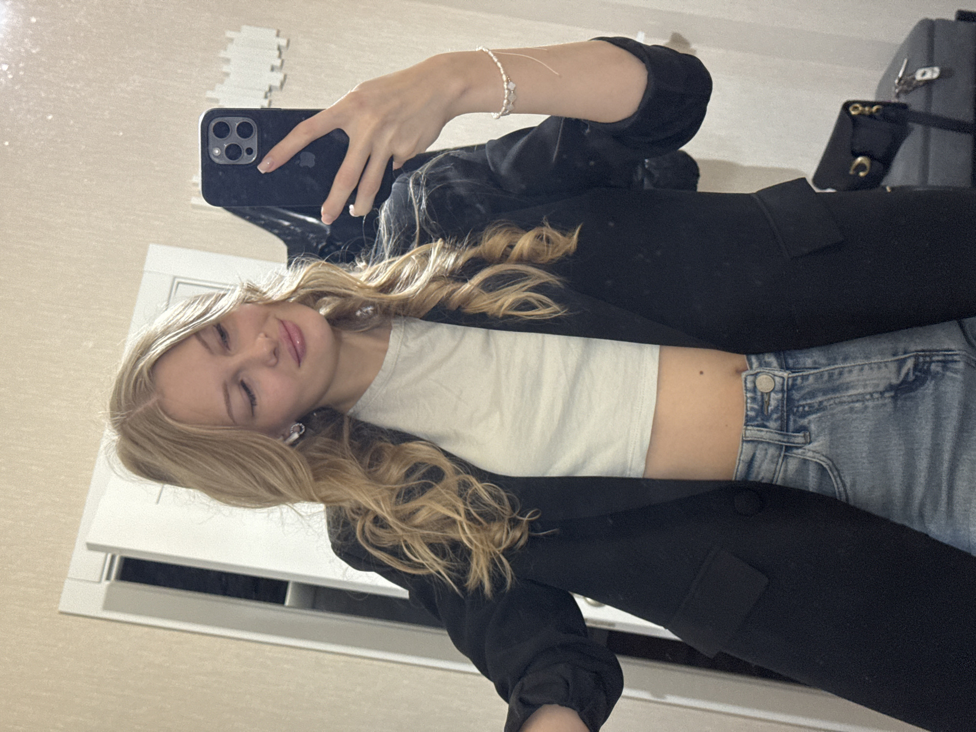 A young blonde woman with light skin, wearing a light-colored crop top, a black blazer, and light-wash denim jeans, takes a mirror selfie. She has on silver stud earrings and a delicate bracelet on her left wrist. Her hair is styled in loose waves. The background appears to be an indoor setting, possibly a hallway or dressing area, with a door and a wall with decorative elements. There is a black purse visible on a gray suitcase in the lower right corner. The image does not contain any elements that can identify a specific location within the city of Perm, Russia.