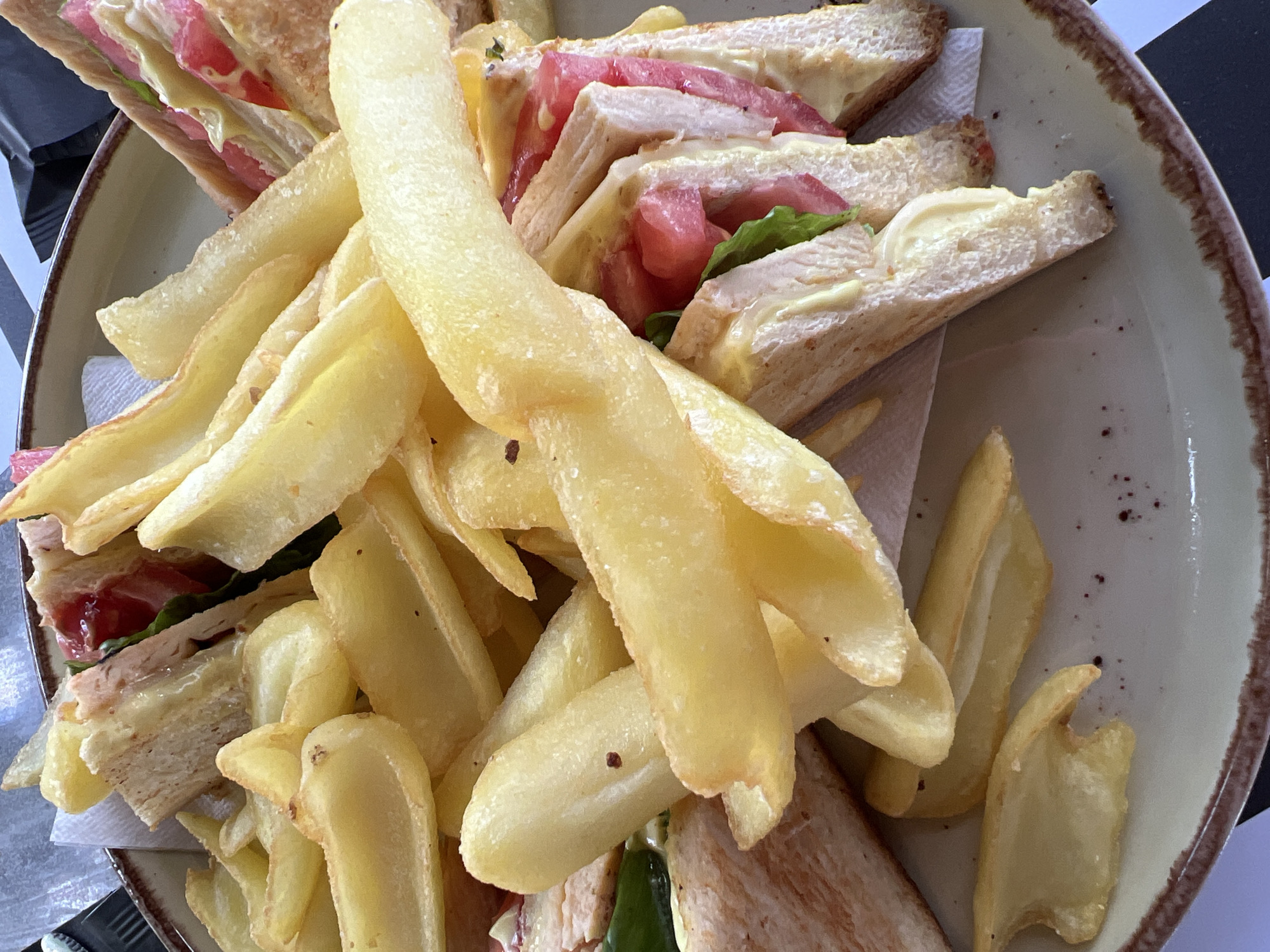 The image shows a close-up, overhead view of a plate of food. The plate contains a serving of french fries and several cut sandwiches. The sandwiches appear to be club sandwiches, with visible layers of bread, meat, lettuce, and tomato. The french fries are thick-cut and golden brown. The food is served on a light-colored plate with a brown rim, which is placed on a surface that appears to be a table. The background is out of focus, but suggests an indoor setting, possibly a restaurant. There is no indication of the specific location within Eretria Municipal Unit, Greece, beyond the fact that this is a food establishment. There are no people visible in the image.