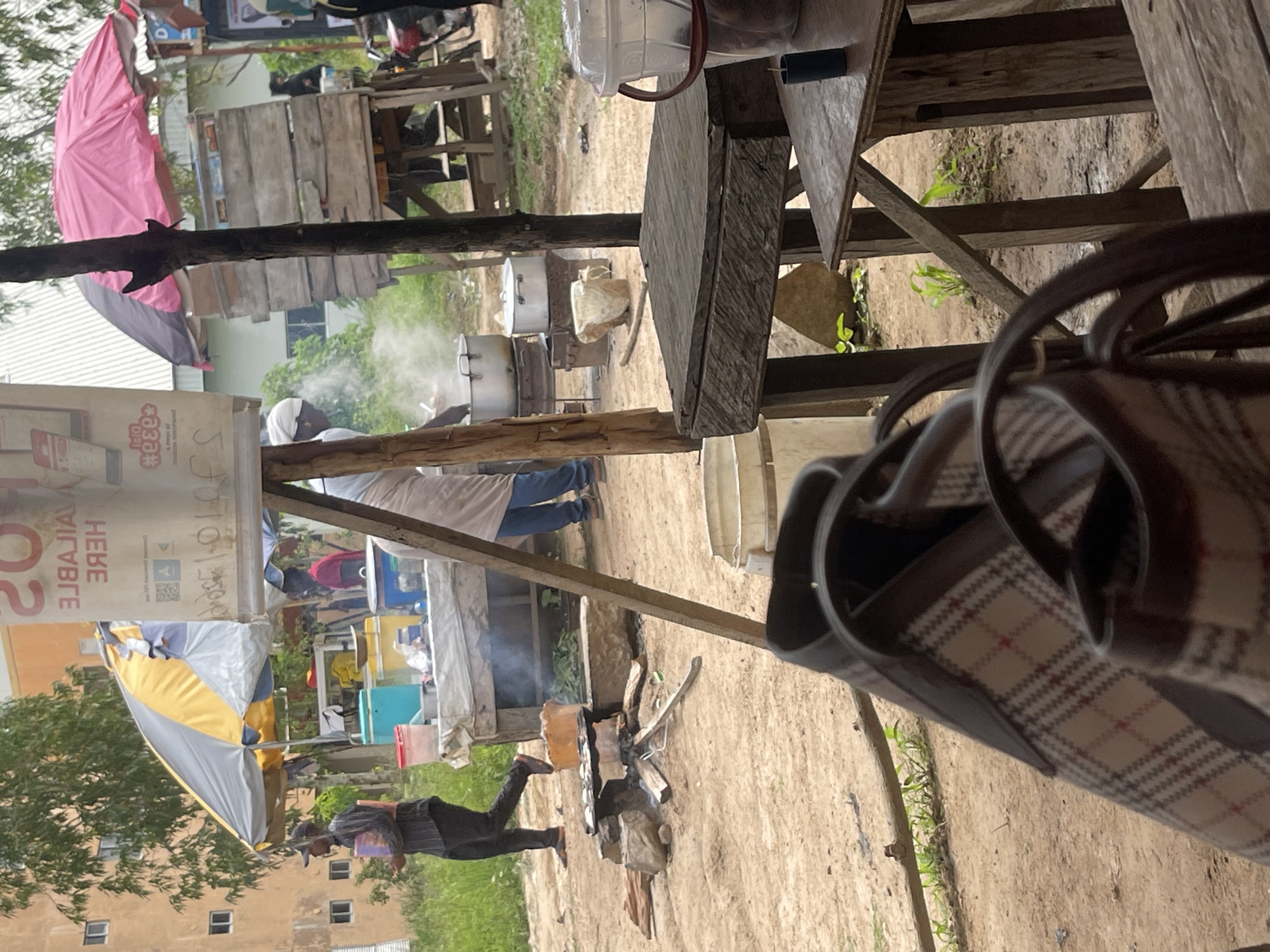 The image depicts an outdoor scene in Rafin Par, Nigeria, likely a street-side food stall or market area. A man in a black striped shirt and black pants is walking to the left, holding a red and purple rectangular object. In the background, a stall with a yellow and gray umbrella is visible, with various items displayed on a table including a teal cooler. Further to the right, another stall with a pink and black umbrella is set up, featuring wooden shelves. Cooking is taking place on an open fire pit with stones and wood. A person in a white apron is tending to large pots emitting steam. Several wooden tables are present in the foreground, one of which has a plaid patterned bag resting on it. The ground is dirt and sand, with some patchy green grass. The overall impression is of a bustling, informal commercial area.