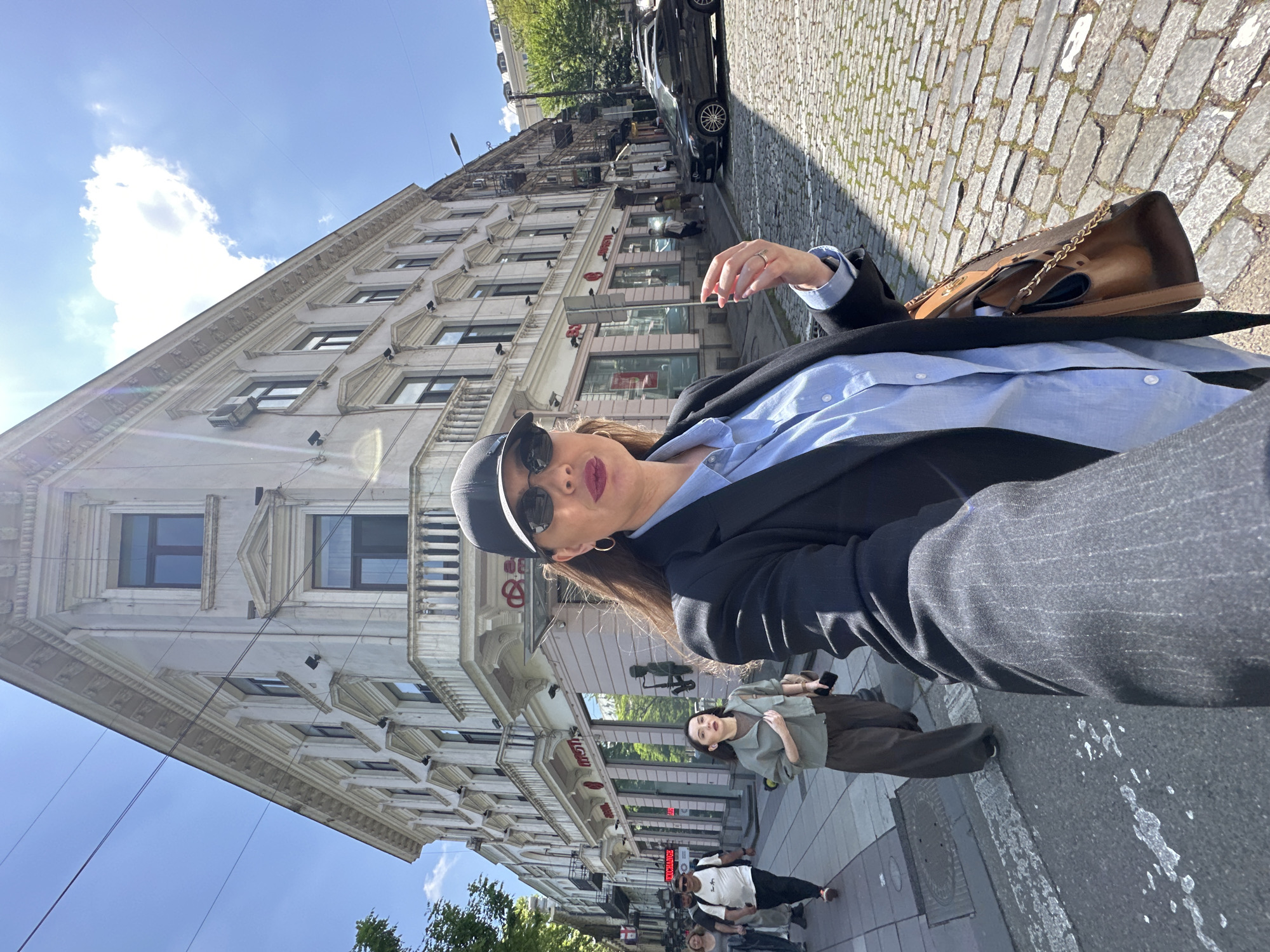 A woman in a blue shirt and black blazer is photographed from a low angle on a cobblestone street. She is wearing sunglasses and a hat, and carrying a brown purse. Behind her is a tall, light-colored building with multiple windows and ornate architectural details. There are shops on the ground floor of the building. Other pedestrians are walking on the sidewalk in the background. A black car is parked on the side of the street. The sky is blue with some clouds. Based on the architectural style and visible shop signage, the location is likely in the city of Tbilisi, Georgia.