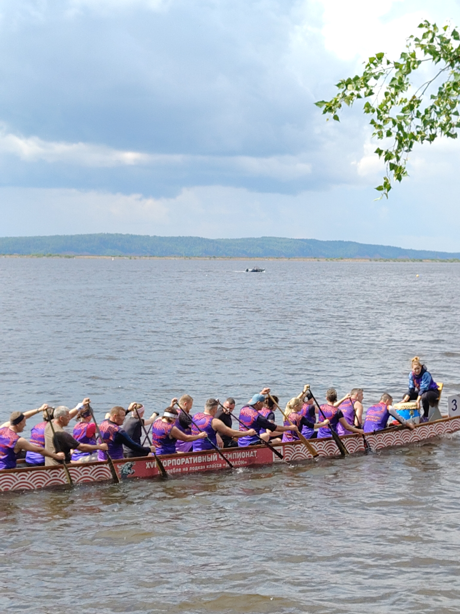 In an outdoor scene at an unspecified location, a dragon boat with a team of paddlers is moving across a body of water under a cloudy sky. The boat is decorated with red and white wave patterns and has text printed on its side. There are approximately fourteen individuals visible, all seated and holding paddles. They appear to be participating in a race or competition, as indicated by their synchronized rowing posture and the race number "3" visible on the back of the boat. One person, likely a coxswain, is seated at the stern of the boat, facing the paddlers, and appears to be steering or directing the team. Another individual, possibly a coach or observer, is also seated at the stern. The background features a distant treeline and a hazy horizon. The water is slightly rippled, suggesting movement from the boat and a gentle breeze. A small boat is visible in the distance on the left side of the frame.