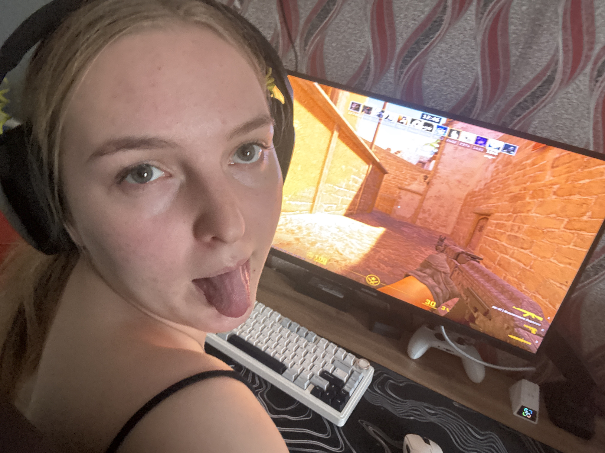 A person with light hair is sticking their tongue out while wearing a black headset. In the background, a computer monitor displays a first-person shooter game with orange brick walls. A white keyboard and a white gaming controller are visible on the desk below the monitor, with a gaming mouse to the left. The wallpaper behind the monitor has a gray and red wavy pattern. Based on the computer's time display of "12:46" and the Cyrillic characters on the screen, the location is likely in Russia. Without additional details, it's impossible to verify the exact location in Perm or otherwise.