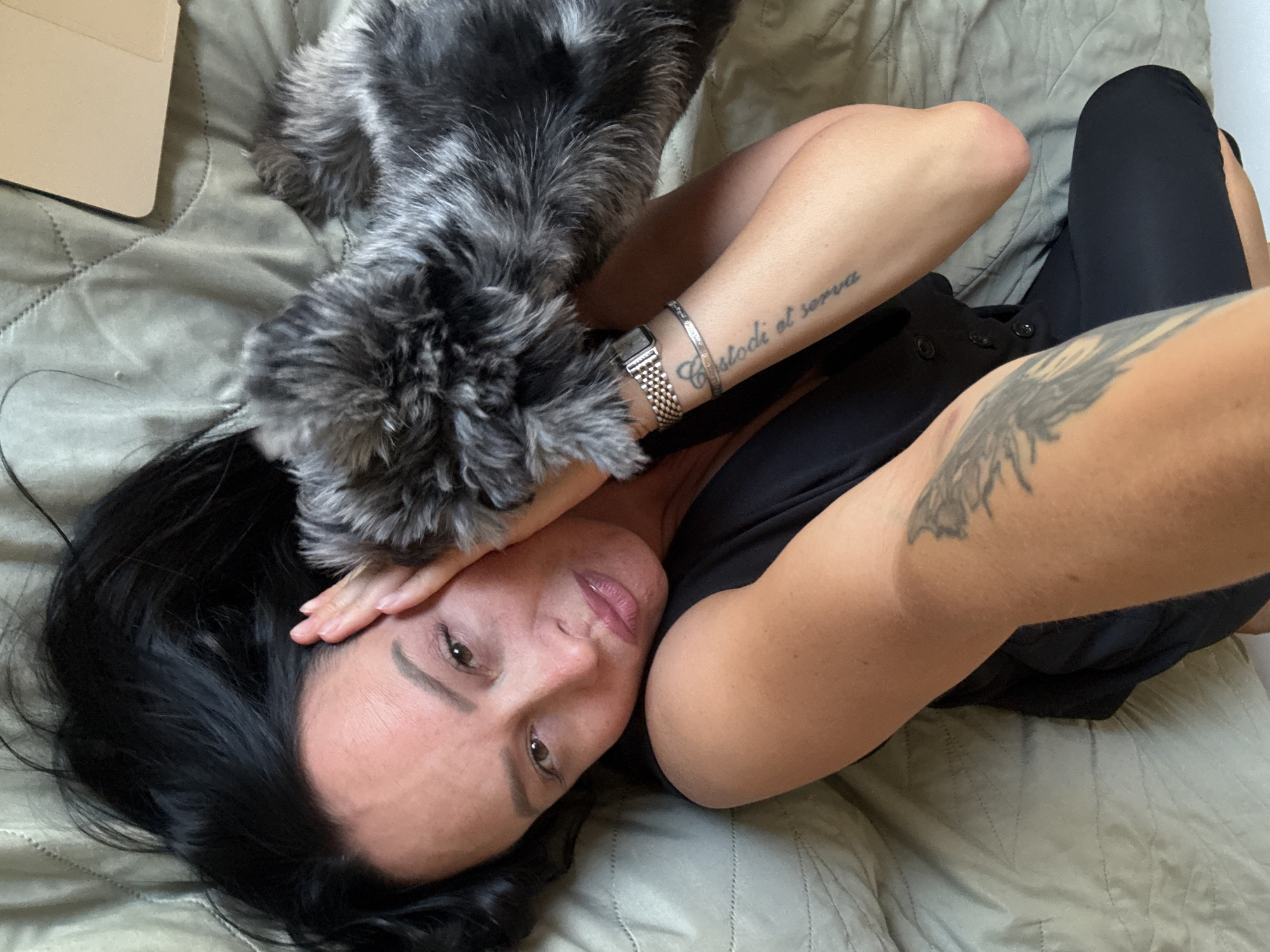 A high-angle, medium shot captures a dark-haired woman lying on her back on a textured, muted green surface, possibly a bed or sofa. She is wearing black clothing with visible buttons down the front. A small, grey and black dog with shaggy fur rests its head near her face, obscuring part of her left cheek and eye. The woman's right arm is bent, supporting her head, with a visible tattoo on her upper arm and forearm, the latter featuring script. A silver watch and bracelet adorn her left wrist. Her face is tilted upwards, eyes open, and her lips are slightly parted, appearing to be wearing lip gloss. The background is a muted green, quilted surface, and a corner of a beige electronic device, possibly a tablet with a cover, is visible in the lower left of the frame. There is no indication of the city of Chaikovsky, Russia, in the image.