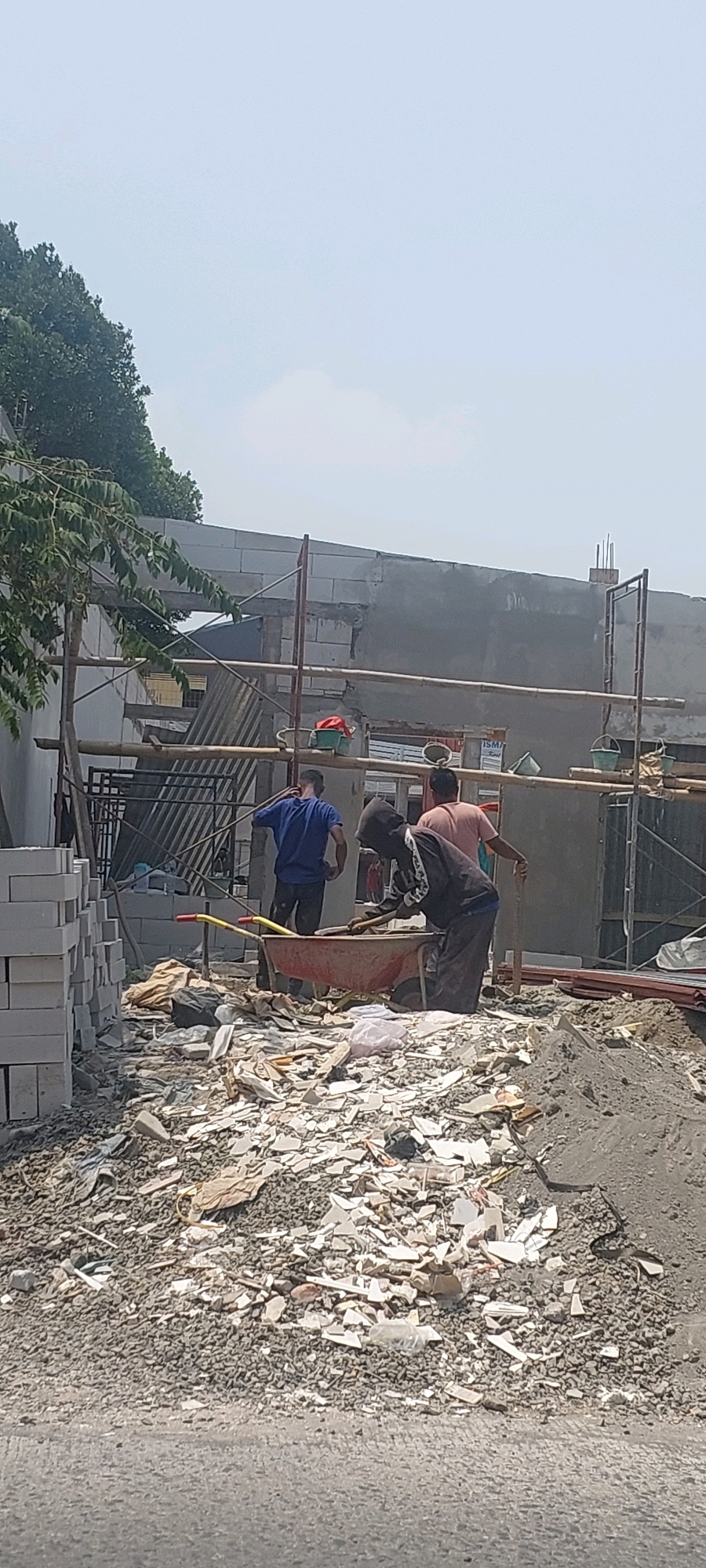 In Semarang, Indonesia, an outdoor construction site is depicted under a clear, hazy sky. The scene is dominated by a large pile of rubble, composed of broken concrete, tiles, and other debris, in the foreground. A stack of white construction blocks is visible to the left. Several individuals are engaged in work. One person, wearing a blue shirt and dark pants, stands near a wheelbarrow. Another, in a dark hooded jacket with white accents and dark pants, is actively working with a shovel in the wheelbarrow. A third individual, in a pink shirt and dark pants, stands nearby, also seemingly involved in the construction activity. Scaffolding, constructed from metal poles and bamboo, is erected along the partially built concrete wall in the background. Various buckets and tools are scattered around the work area. The overall impression is one of ongoing construction and debris removal.