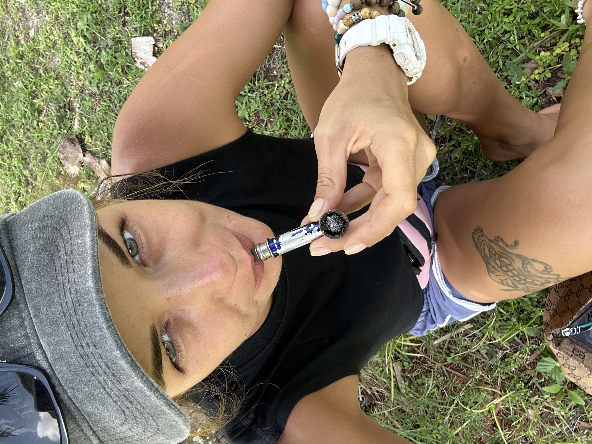 The image shows a close-up of a person sitting on the grass. The person is wearing a black t-shirt and blue and pink shorts. They are also wearing a grey baseball cap and a white watch with multiple beaded bracelets on their wrist. The person is holding a pipe with what appears to be tobacco or marijuana up to their lips. A tattoo of a dragon is visible on their thigh. A designer bag with a monogram pattern is lying next to them on the grass. The background consists of green grass and some dry leaves. The lighting suggests it is daytime, and the overall setting appears to be outdoors in a natural environment. The person's face is partially visible, and they are looking towards the camera with a neutral expression. There is no visible text in the image.
