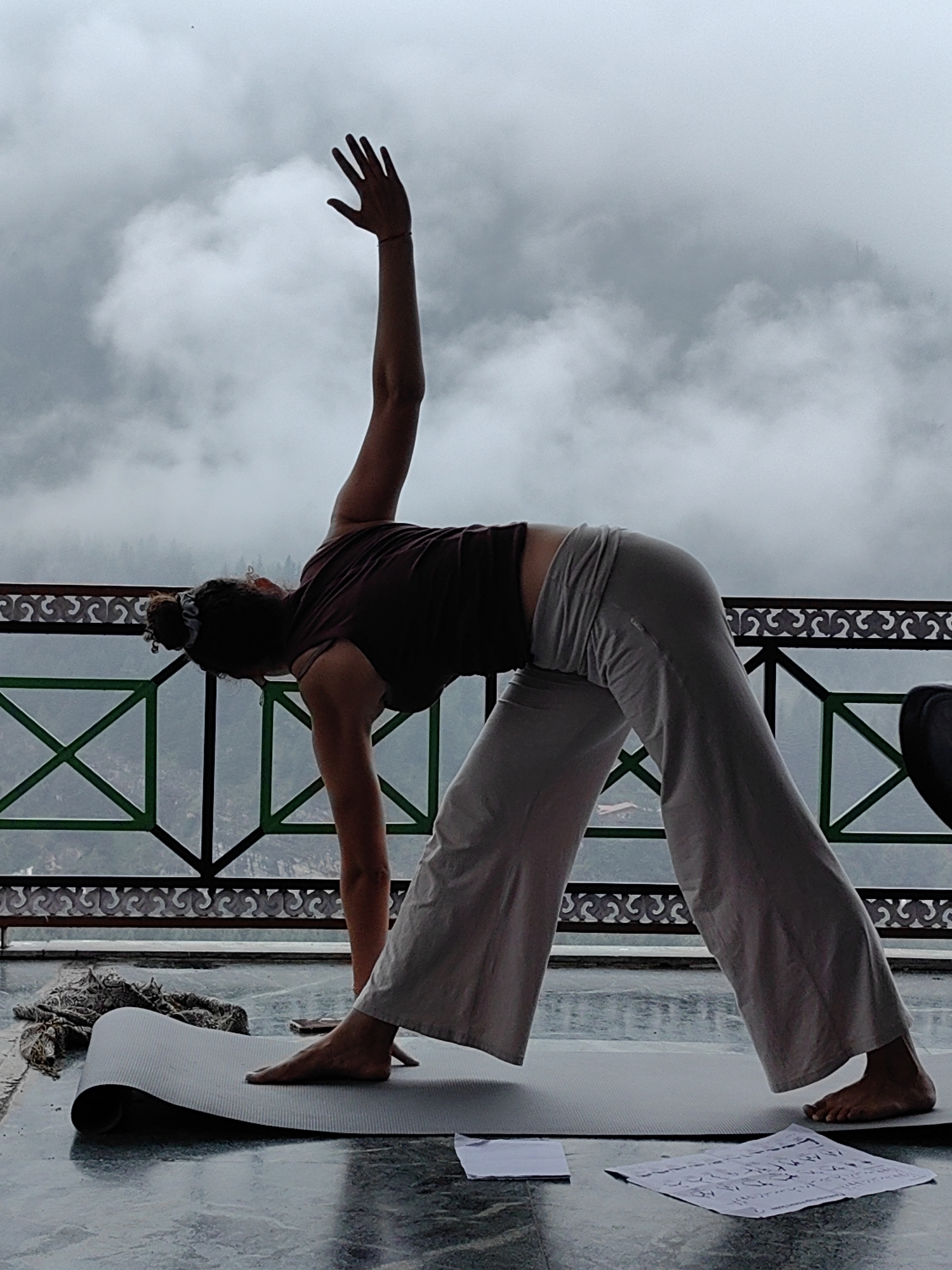 A woman performs a yoga pose on a balcony in Vashisht, India. She is wearing a dark maroon tank top and loose-fitting grey pants. Her right arm is extended upwards, and her left hand is placed on her left leg. Her left foot is planted firmly on the ground, while her right leg is extended backward. The woman is on a grey yoga mat, which is placed on a tiled floor. In front of the mat are two pieces of paper with writing on them. A patterned scarf lies to the left of the mat. Behind the woman, a black metal railing with decorative white accents separates the balcony from a misty, mountainous landscape. The sky is overcast with thick clouds.