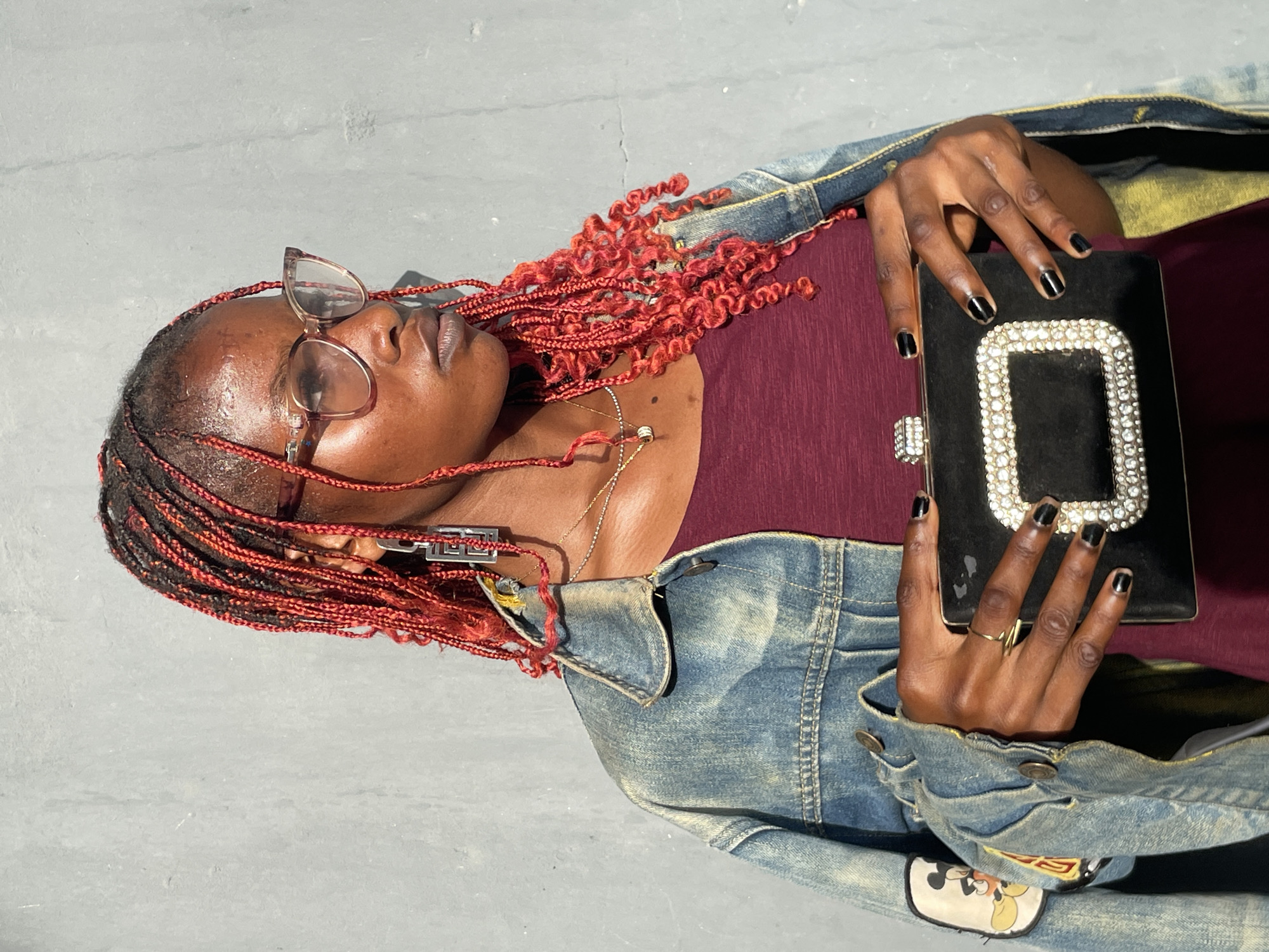 The image features a dark-skinned individual, likely female, captured in a brightly lit outdoor setting against a plain, light gray background, possibly concrete or pavement, in Badagry, Nigeria. The subject is shown from the chest up, with her head tilted and looking slightly upwards and to the side, wearing light-framed glasses.
Her hair is styled in long, vibrant red braids, with some strands appearing curly or wavy at the ends. Her skin shows a slight sheen, possibly due to sweat or oil, suggesting warm or humid weather. She is dressed in a maroon or burgundy top, layered with a light blue, distressed denim jacket. A notable detail on the jacket is a colorful patch featuring the character Mickey Mouse, visible on the lower left arm area.
She holds a black rectangular clutch purse with both hands, positioned in front of her. The clutch is decorated with a prominent, sparkling silver embellishment that forms a large rectangular outline on its front surface. Her fingernails are painted with black nail polish, and a simple gold ring is visible on her right ring finger. A delicate gold chain necklace is also visible around her neck. The image appears to be a posed shot, capturing a casual yet accessorized look.