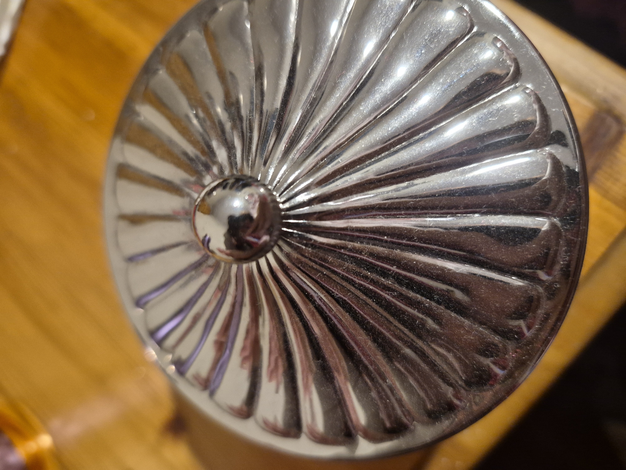 A close-up, overhead shot shows a reflective, fluted silver lid with a spherical knob in the center, resting on a wooden surface. The lid has a radial pattern of raised ridges. The wooden surface appears to be a table with a warm, light brown tone and visible grain. There is no discernible location in the city of Sandwell, United Kingdom.