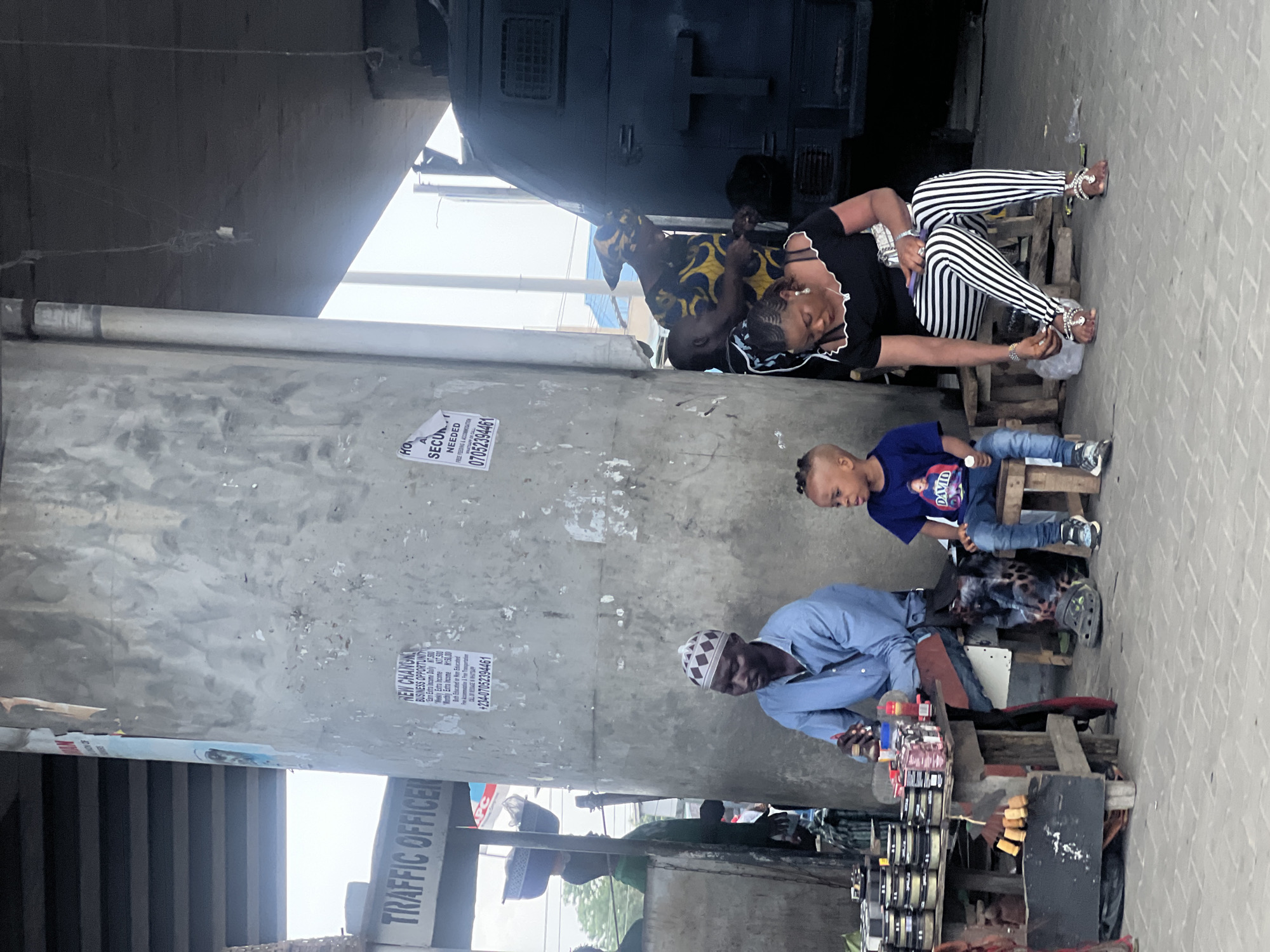 The image captures a scene in Ado, Nigeria, showing individuals engaged in various activities under an overpass. In the foreground, an adult male, wearing a light blue shirt and a patterned white cap, sits on a wooden stool, likely tending to items for sale displayed on a makeshift wooden table. Next to him, a young child in a blue shirt and jeans sits on another wooden stool. Behind them, a woman wearing black and white striped pants and a black top is seated, possibly interacting with another adult. Various objects and printed posters are affixed to the overpass support, including "TRAFFIC OFFICER" on a sign. The setting suggests an urban area. A vehicle of some kind sits parked on the overpass above.