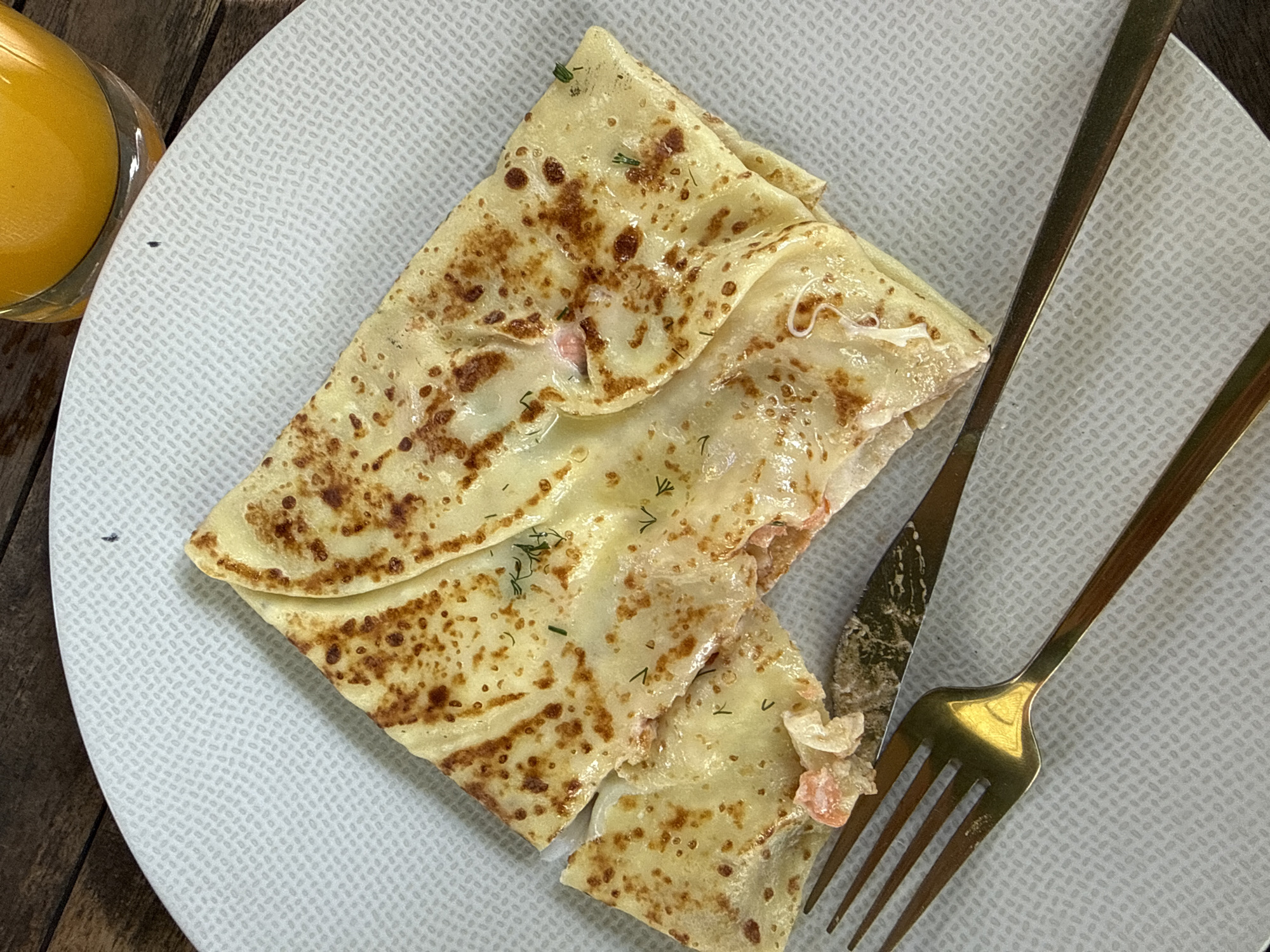 On a white patterned plate, likely indoors on a wooden table, sits a folded crepe filled with what appears to be salmon and dill. To the right of the crepe lies a golden fork and knife. A glass of orange juice is partially visible in the upper left corner of the frame. The location is not possible to determine from the image.