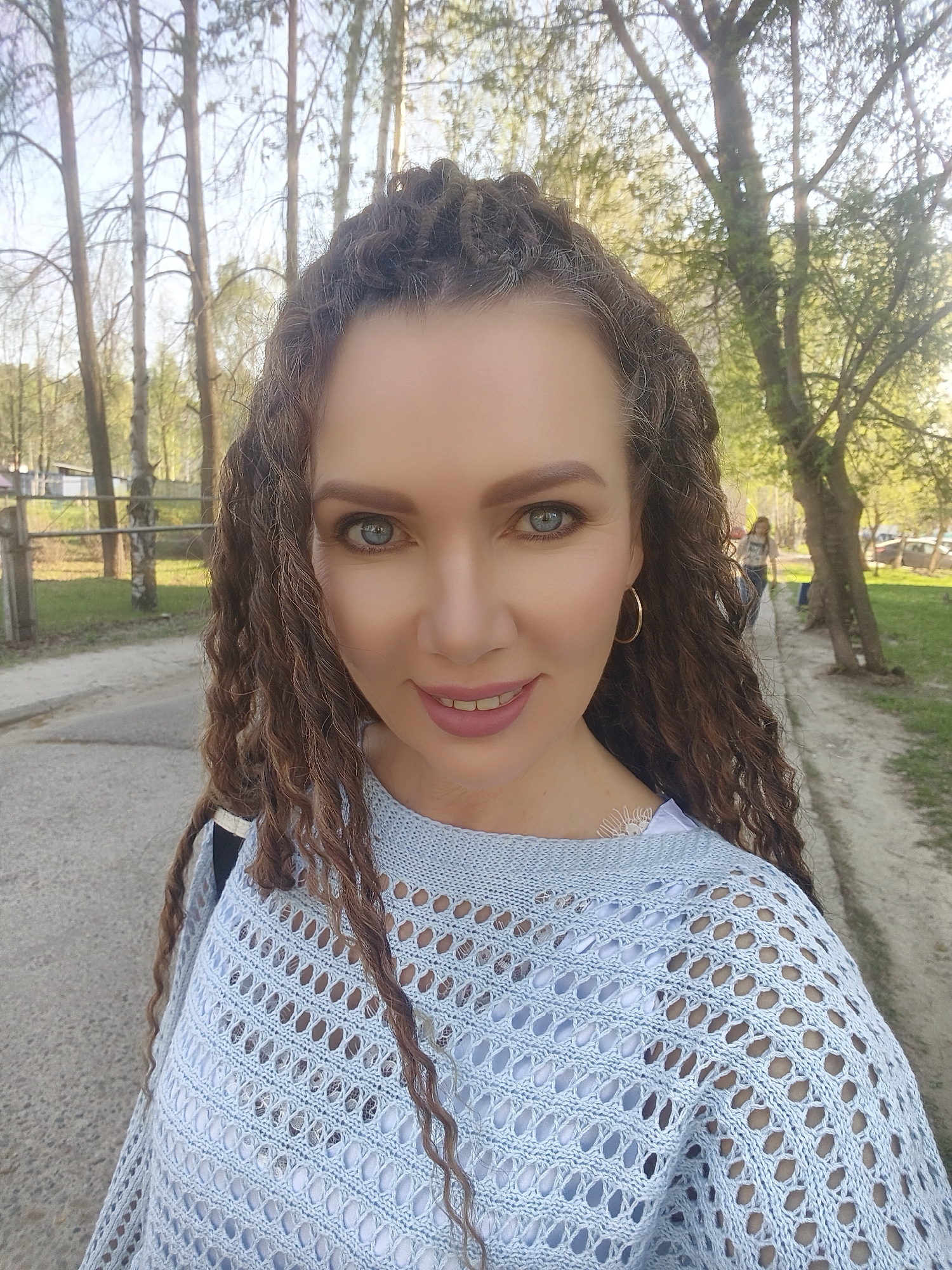 The image shows a woman with curly brown hair, blue eyes, and fair skin. She is wearing a light blue, open-knit sweater over a white top, and gold hoop earrings. The background suggests an outdoor setting, likely a park or tree-lined street, with a path or road visible. There are trees with sparse foliage, indicating it might be early spring or late autumn. Other people are faintly visible in the background, suggesting a public space. The photo appears to be taken in Unknown, Unknown.