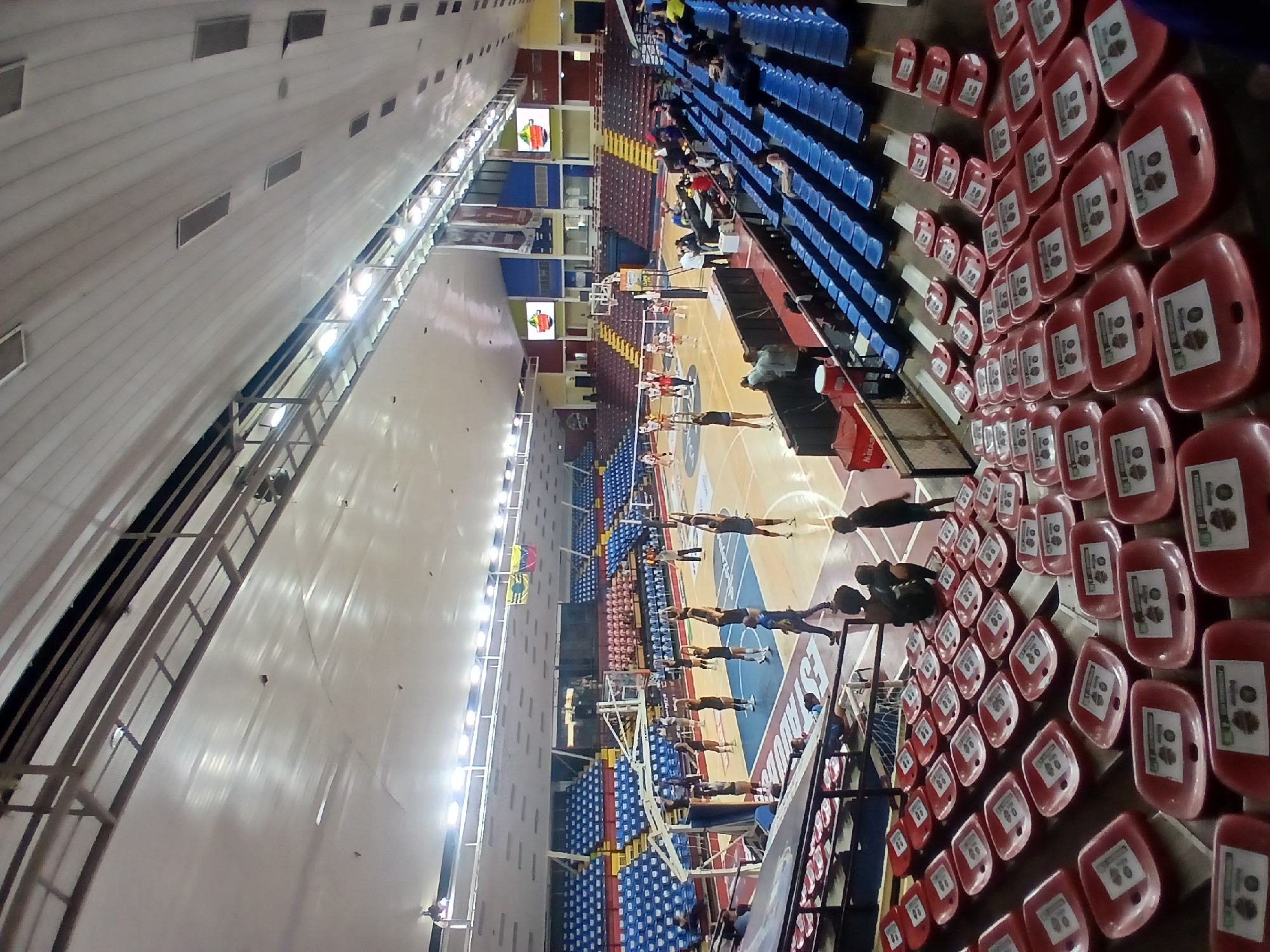 The image shows an indoor volleyball court in Ciudad Guayana, Venezuela, likely inside the CTE Cachamay. A volleyball team practices on the court. Spectator seating, a scoreboard, and the building's interior structure are visible.