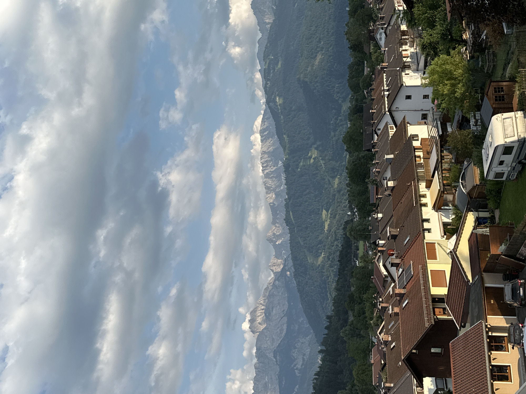 The image captures a panoramic view of a residential area nestled against a backdrop of majestic mountains, under a sky filled with dramatic clouds. The scene appears to be set in Garmisch-Partenkirchen, Germany, as indicated by the architectural style of the houses and the prominent mountain range.
In the foreground, a collection of houses with steeply pitched, red-tiled roofs forms a dense neighborhood. Many of the houses have multiple stories, with visible windows and balconies. Some have solar panels installed on their roofs, suggesting a modern approach to energy. Among the houses, there are trees providing greenery, and some sheds and other structures. A white caravan is parked on the right side of the frame, near a wooden shed. Cars are visible on a driveway.
The middle ground is dominated by a lush, forested mountain slope that rises steeply behind the houses. The trees are dense and green, indicating a temperate climate and likely summer or early autumn.
Further in the background, a rugged mountain range stands tall. The peaks are sharp and rocky, with some snow or white patches visible on the higher elevations. The mountains are partially obscured by a cloudy sky.
The sky itself is a significant element of the image, filled with large, billowy clouds that cast shadows and create a dynamic interplay of light and dark. The clouds suggest a somewhat overcast or rapidly changing weather pattern, possibly late afternoon or early evening given the lighting on the mountains. The overall atmosphere is serene and picturesque, highlighting the harmonious coexistence of human settlement and the natural grandeur of the Alps. No visible text is present in the image.