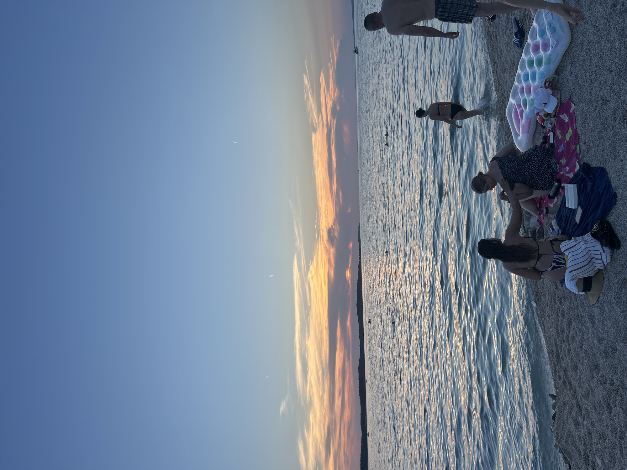 This image captures a group of people at the beach during sunset. The sky is a clear blue with streaks of orange and yellow from the setting sun, indicating it is dusk. The water is calm with gentle ripples reflecting the sky's colors. Several individuals are visible: a person standing in the water, another sitting on the shore next to various beach items like towels and an inflatable raft, and another person further down the beach near the water's edge. The scene appears relaxed and idyllic, suggesting a leisurely summer evening by the sea. The location is a beach with a pebbly shore, and the presence of a raft and towels indicates recreational activity. There is no visible text in the image.
