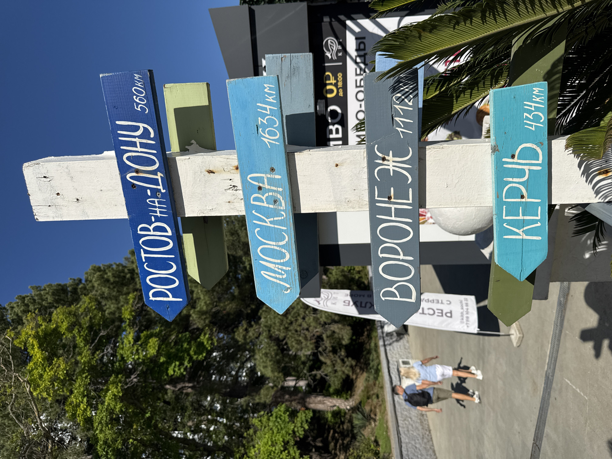 The image captures a vibrant wooden signpost under a clear blue sky, likely in a sunny location such as Sochi, Russia. The signpost, made of a white central column, features four colorful directional planks pointing to different cities and their distances.
From left to right (as oriented in the main image), the signs read:
1. A dark blue sign: "РОСТОВ-НА-ДОНУ 560км" (Rostov-on-Don 560 km).
2. A light blue sign: "МОСКВА 1634 км" (Moscow 1634 km).
3. A grey-blue sign: "ВОРОНЕЖ 1112" (Voronezh 1112), likely implying kilometers.
4. A turquoise sign: "КЕРЧЬ 434 КМ" (Kerch 434 KM).
The scene is set outdoors on a bright, sunny day, evidenced by the strong shadows and clear weather. Lush green trees, including some palm-like foliage, suggest a warm climate. In the background, two people, a man and a woman in casual attire, are seen walking away from the viewer on a paved area. Behind them, parts of modern buildings with dark facades and white elements are visible, along with a white banner.
Further visible text on the buildings and banners includes "ВО UP" and "до 18:00" (until 18:00), possibly indicating business hours. Another sign reads "МНО-ОБЕДЫ" (MNO-LUNCHES or MNO-DINNERS). A white banner displays "РЕСТ СТЕPPA" (likely "Restaurant Terra") along with a phone number: "7318 104 6022". The overall impression is a lively resort or public area.