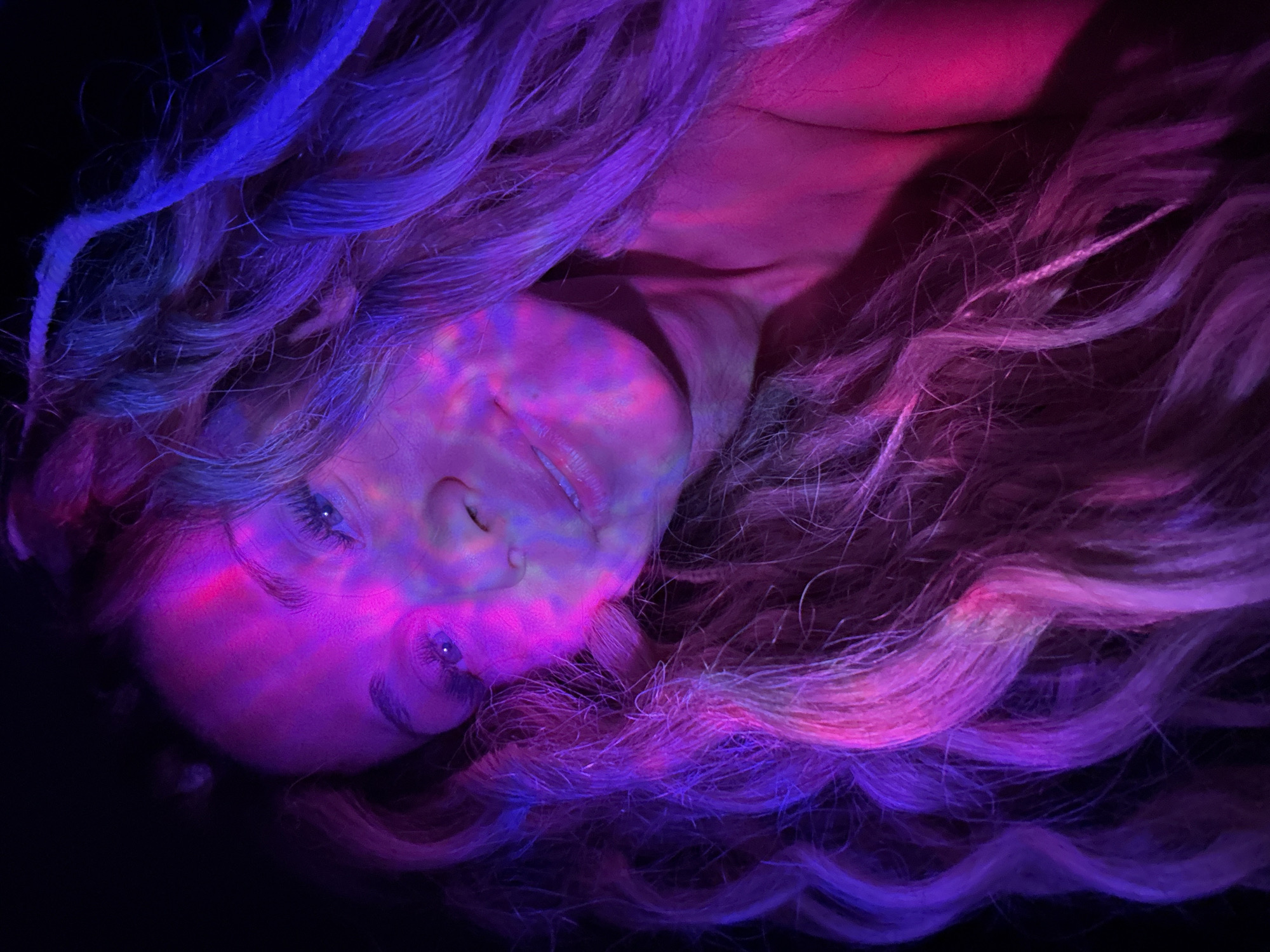 A low-angle close-up photograph depicts a woman with long, wavy blonde hair resting against a dark background. Her face is illuminated with a mixture of pink and blue light, creating a patterned effect. Her eyes are partially closed, and her expression is serene. The hair is spread out around her head, exhibiting variations in color from blonde to purple under the lighting. No city or location information is discernible from the image.