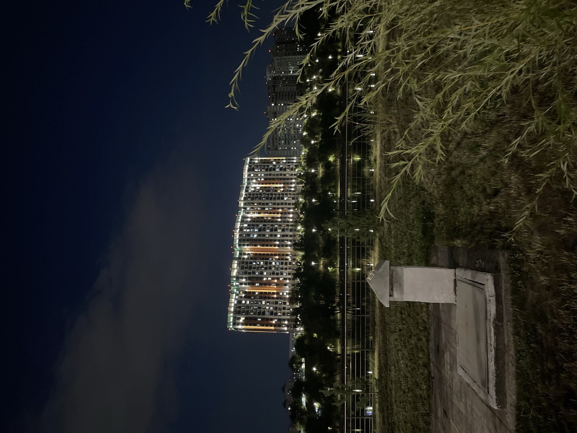 The image depicts a nighttime scene in Almaty, Kazakhstan, likely in a park or recreational area adjacent to a body of water. In the background, a large, modern residential building is illuminated with numerous lights, suggesting it is a multi-story complex. The building has a distinctive pattern of lights, with some sections featuring orange vertical stripes. In front of the building, there is a line of trees and streetlights bordering a body of water, possibly a river or a lake, which reflects the lights. The foreground of the image shows a grassy area with some dry vegetation and foliage from trees or bushes encroaching into the frame from the right. A concrete structure with a square cover and a small, angled roof-like cap is visible in the lower left quadrant, appearing to be some form of utility access or monument. The sky is dark and shows some faint clouds.