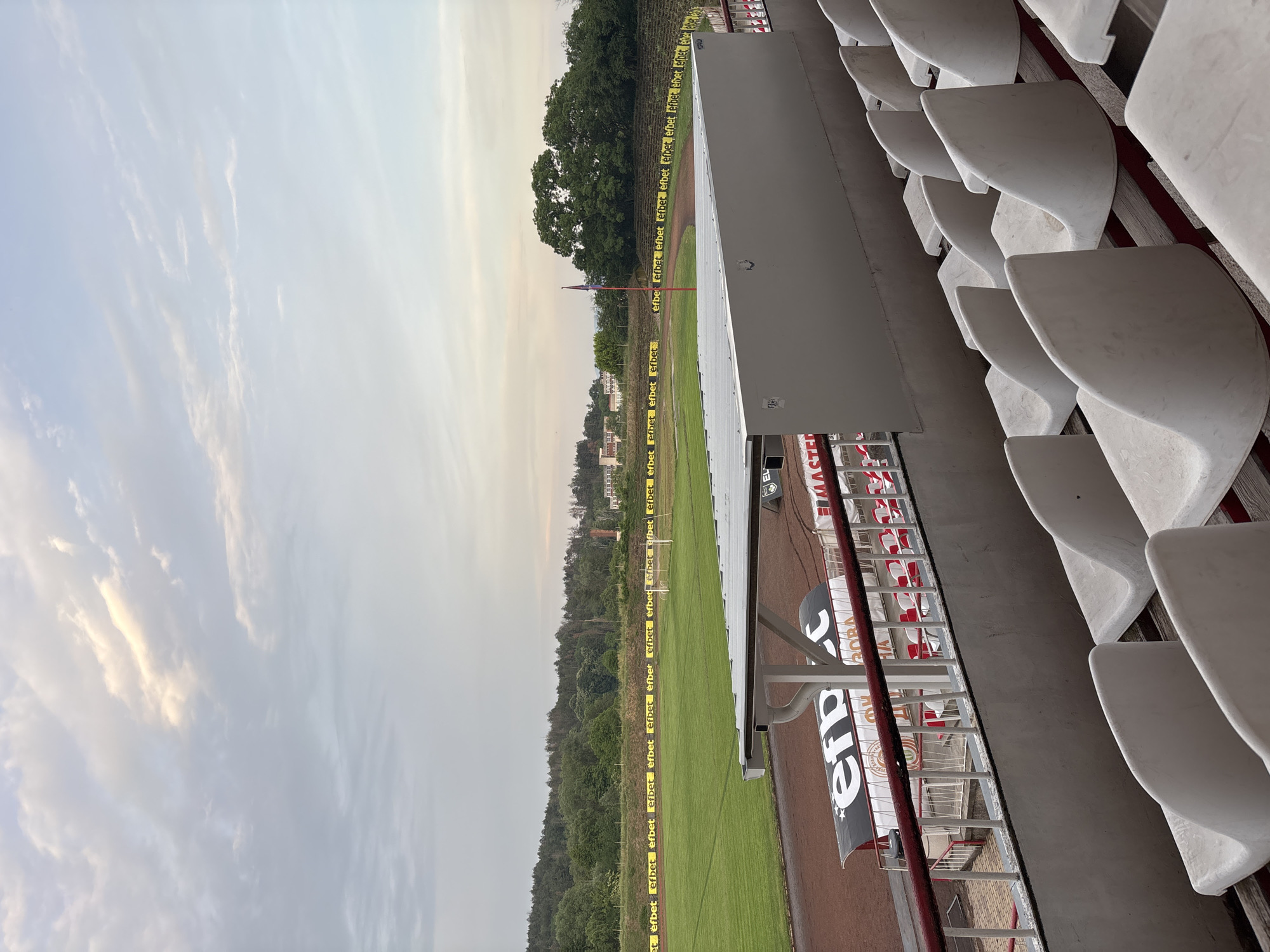 This image presents a wide-angle view of an empty sports stadium, likely a football pitch, captured from the perspective of the stands. The scene is set under an overcast sky, suggesting late afternoon or early evening with soft, diffused lighting.
In the immediate foreground on the right, several rows of white, unoccupied stadium seats are visible. Below these stands, there is a lower section with supporting structures and banners. Visible text on these banners includes "BMASTER" and "efpc" on white and black backgrounds with red accents.
The main focus of the image is the green grass playing field, surrounded by a reddish-brown running track. Along the perimeter of the field, numerous yellow advertising hoardings conspicuously display the word "efbet" multiple times, a prominent Bulgarian betting company, consistent with the location context of Kazanlak, Bulgaria. A goalpost is visible at the far end of the field, and a flagpole with a dark flag stands near the track.
Beyond the stadium, a dense line of lush green trees forms the backdrop, with some distant buildings partially discernible through the foliage. The sky above is largely cloudy with soft white and light grey tones. There is no activity taking place, and no people are present, lending a quiet, still atmosphere to the scene.